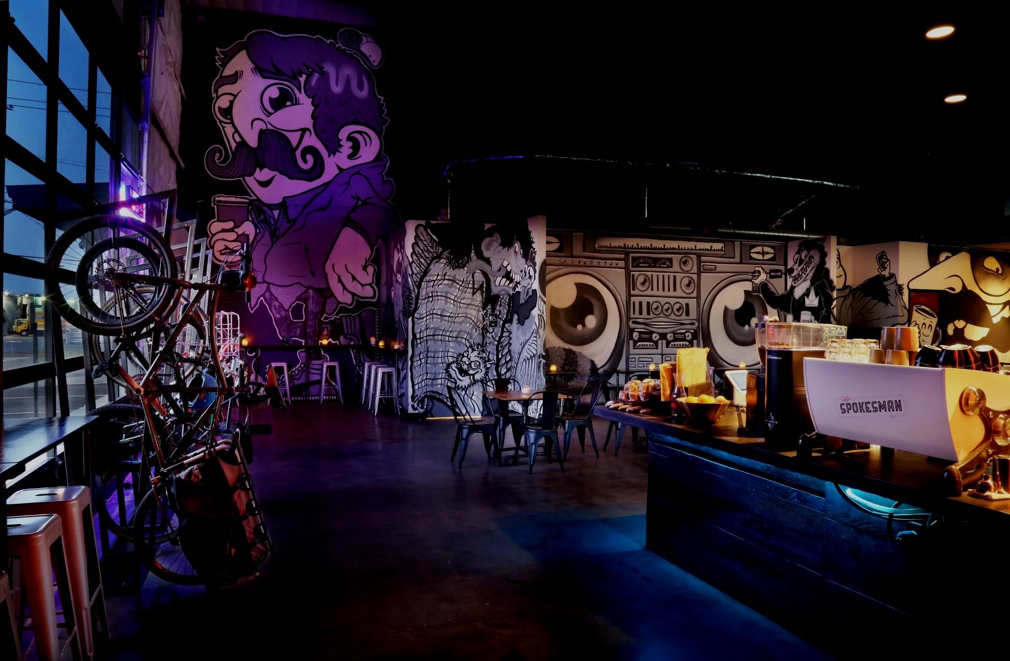 Interior of a cafe with graffiti art on the walls, including characters with exaggerated expressions. Bicycles are displayed on a rack near a large window, and there are tables with chairs and bowls of food or snacks. The space is dimly lit with some ceiling lights.