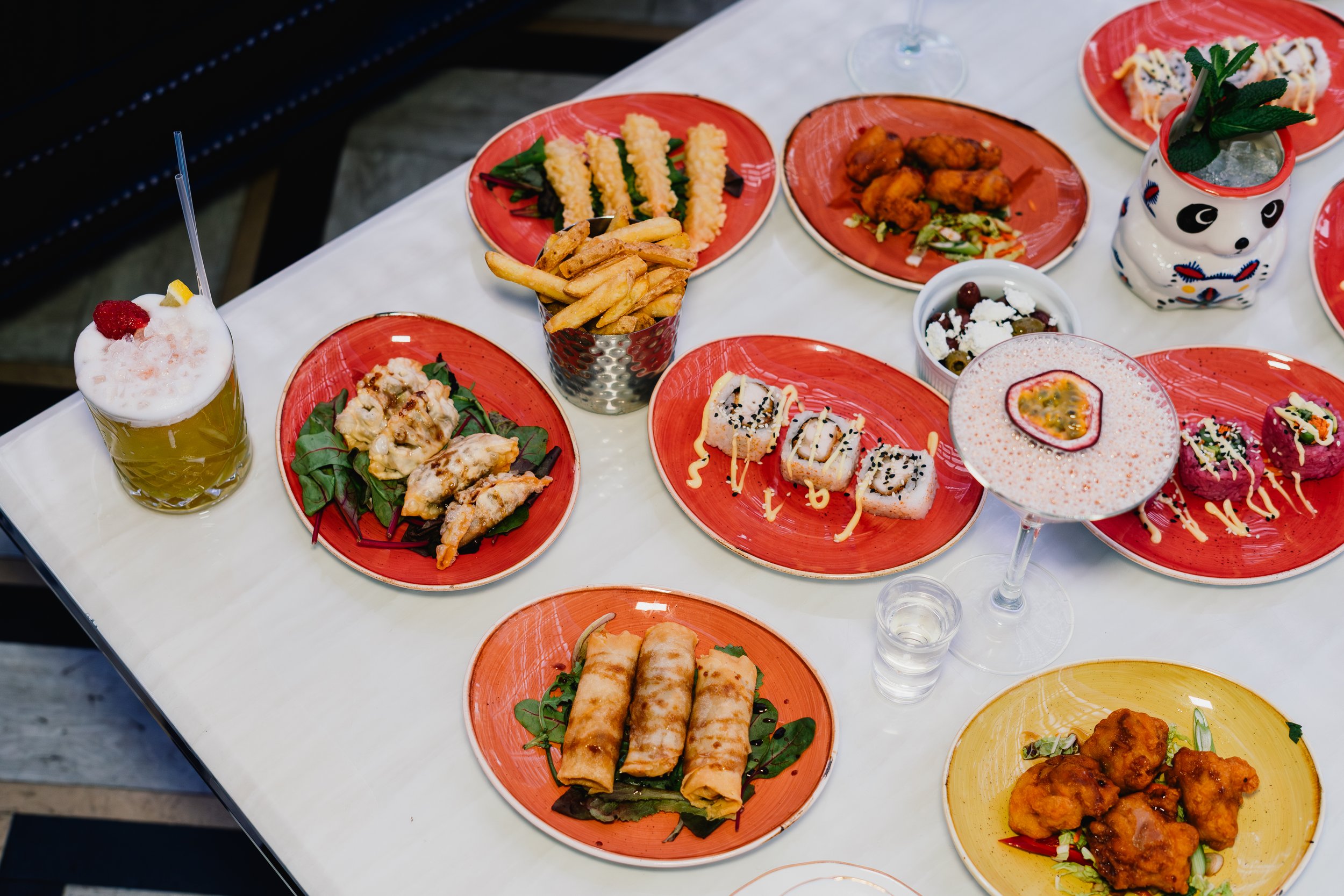 A table with various plates of Asian cuisine including sushi, spring rolls, fried chicken, French fries, and desserts, with drinks and a panda-shaped container with mint and a straw.