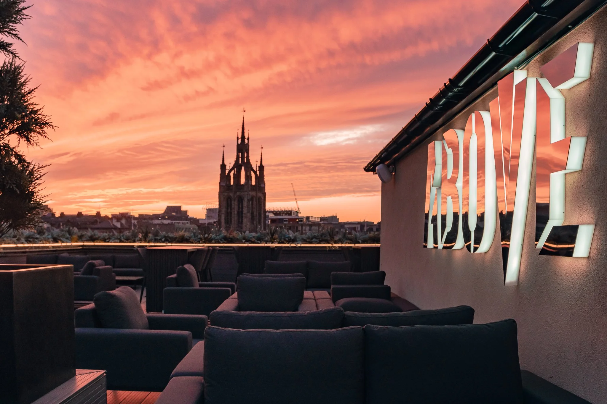A rooftop bar overlooking a city with a sunset sky, a tall church spire, and modern seating and a neon sign reading 'ARCERY' on a wall.