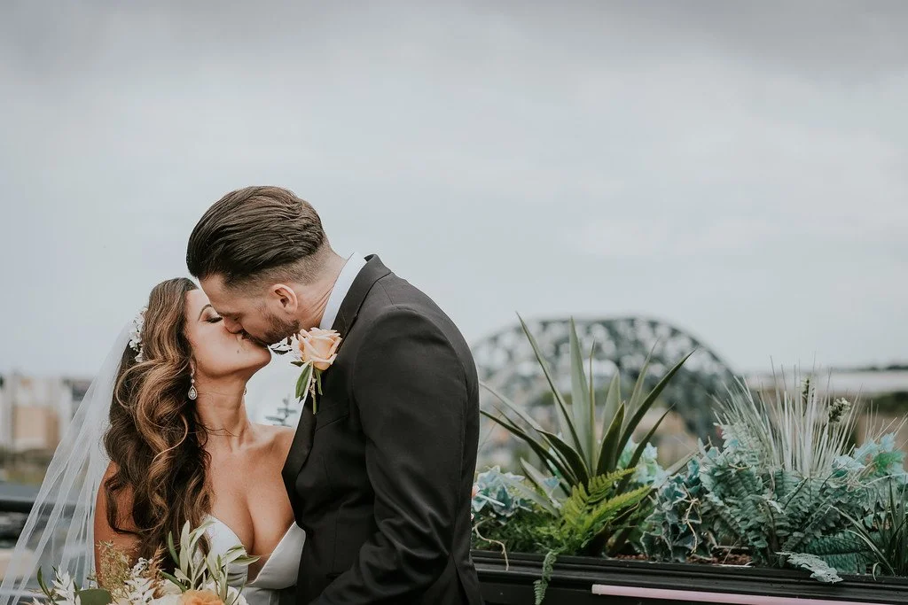 A bride and groom kissing outdoors on a cloudy day, with a bridge in the background and greenery around them.