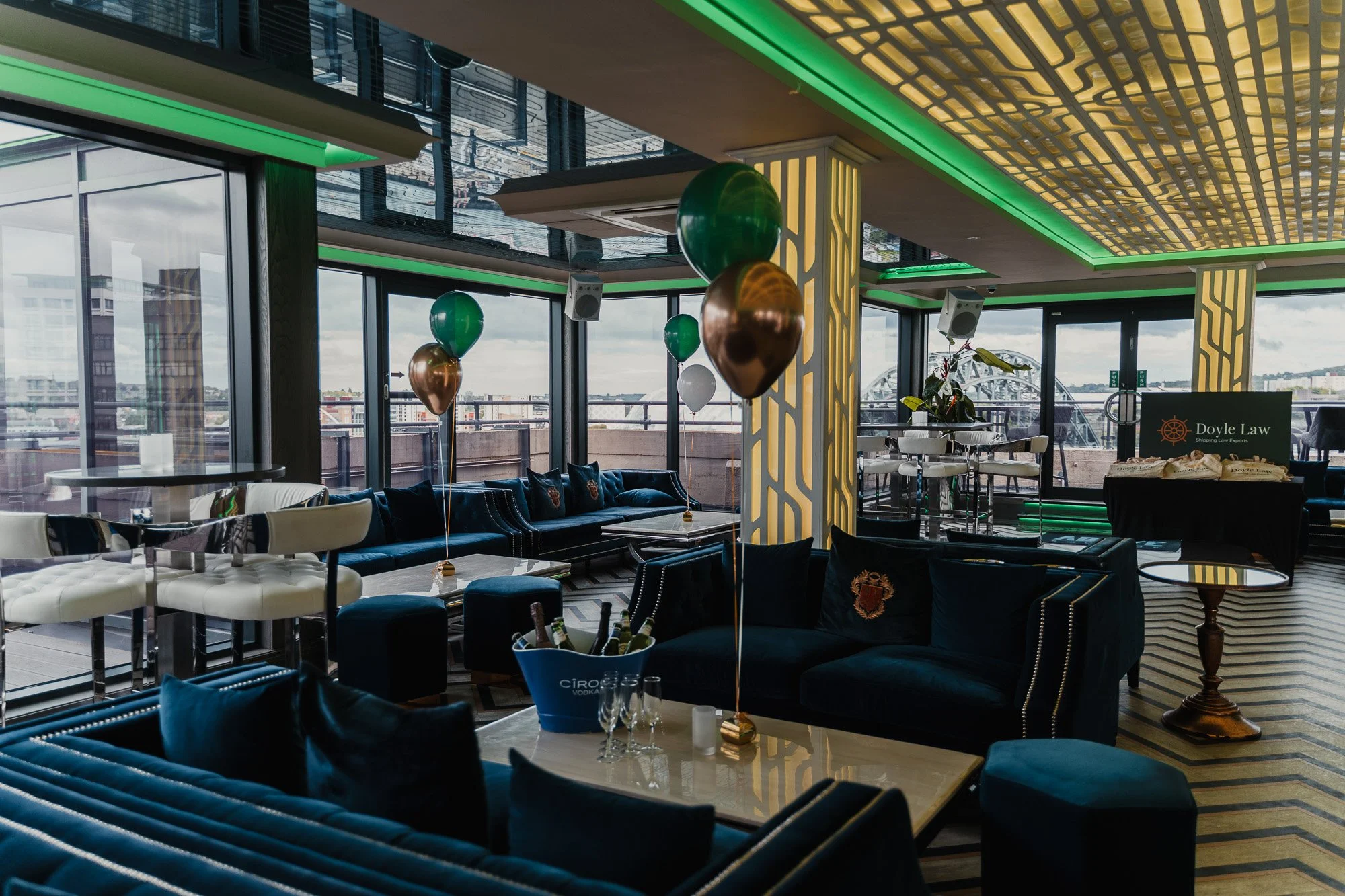 Interior of a modern upscale lounge or restaurant with large windows, colorful balloons, and stylish furniture, including cream and dark blue chairs and white tables, decorated for a celebration.