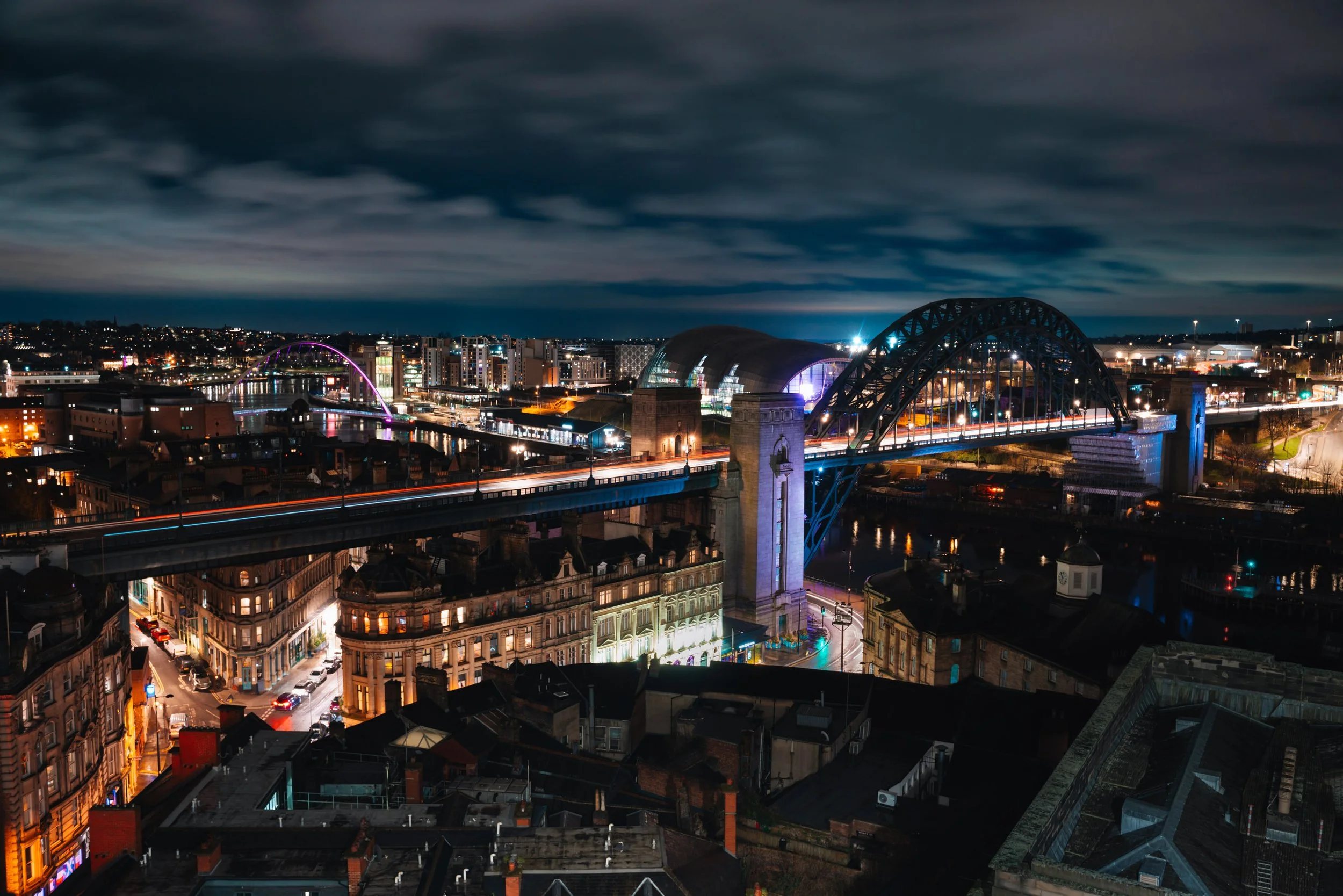 Nighttime cityscape with illuminated bridges, colorful lights, and dark cloudy sky.