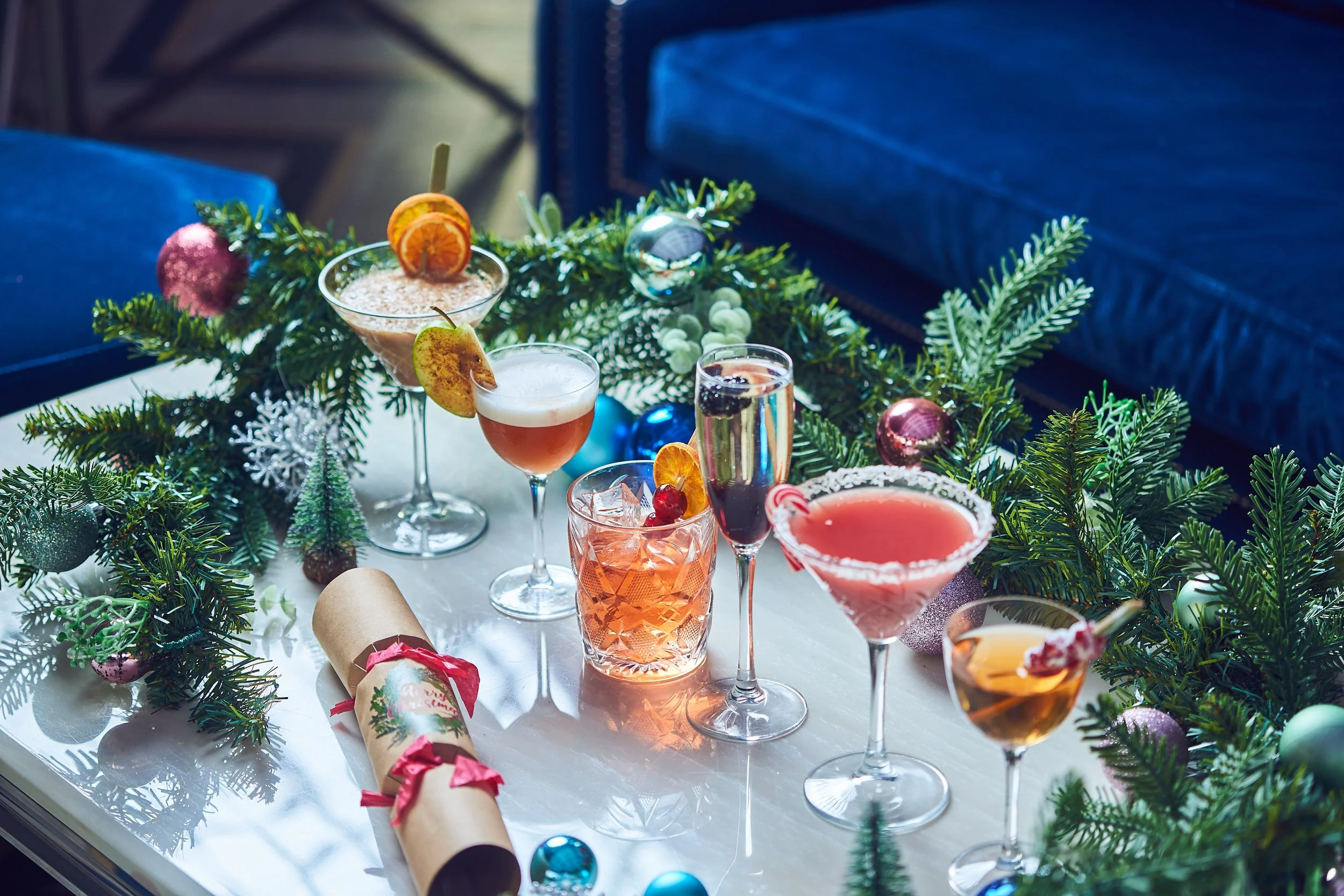 A festive holiday table decorated with green pine branches and colorful ornaments, featuring various cocktails in fancy glasses, some garnished with fruit slices, surrounded by Christmas-themed decorations and confetti.