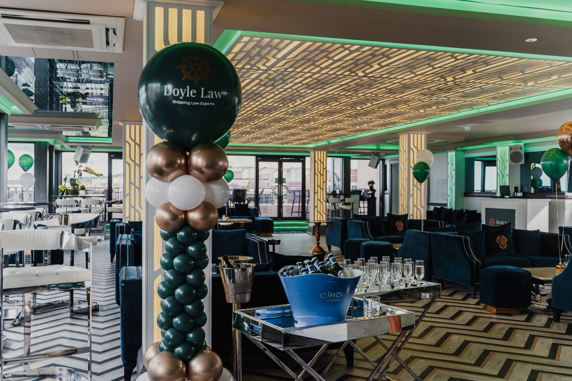 Inside a decorated event space or restaurant with balloons, tables, glasses, and modern decor, featuring a balloon column with a Doyle Law sign and a geometric ceiling design.