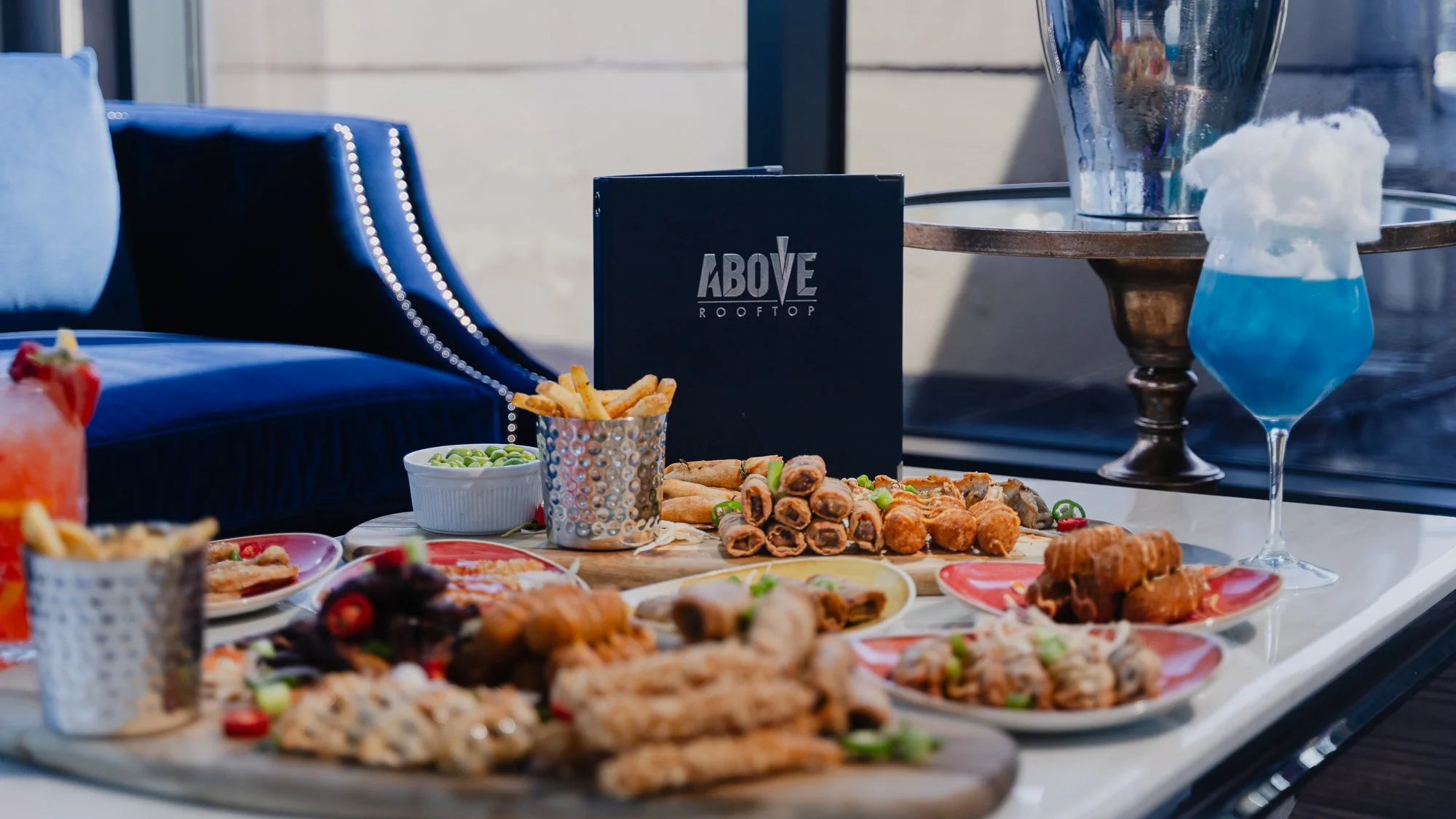 A table with various appetizers and snacks, including spring rolls, fried chicken, and other finger foods, set outside with a sign reading 'Above Rooftop' in the background.