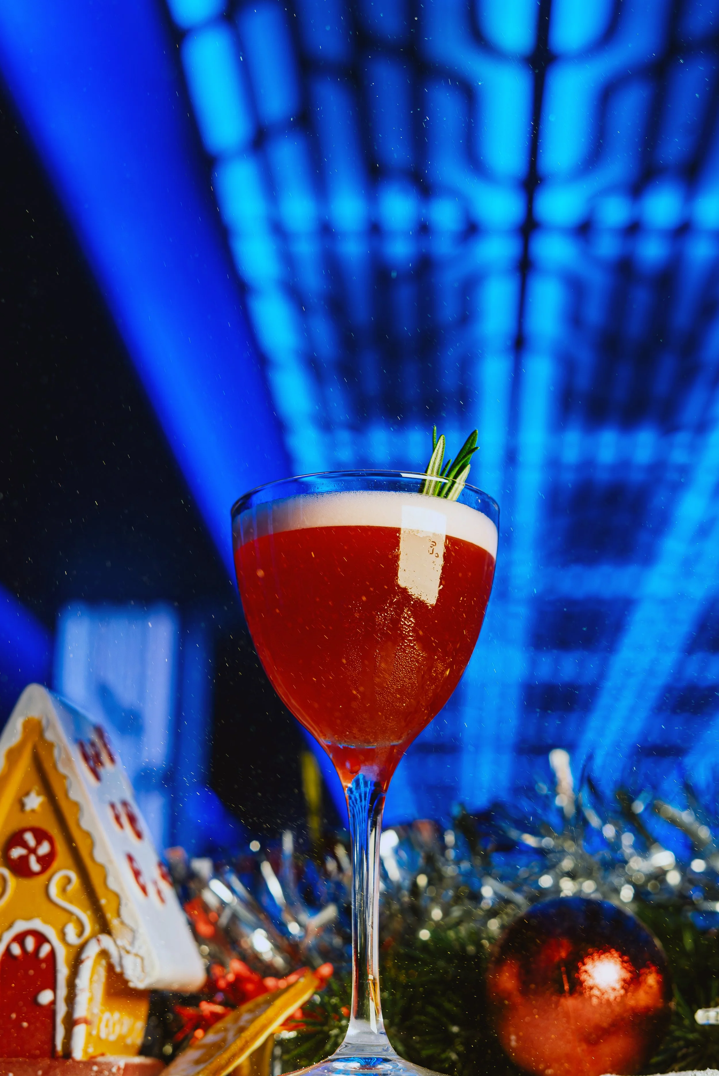 A cocktail glass filled with a red drink and garnished with herbs, set against a blurred festive background with blue lighting and bokeh effects.