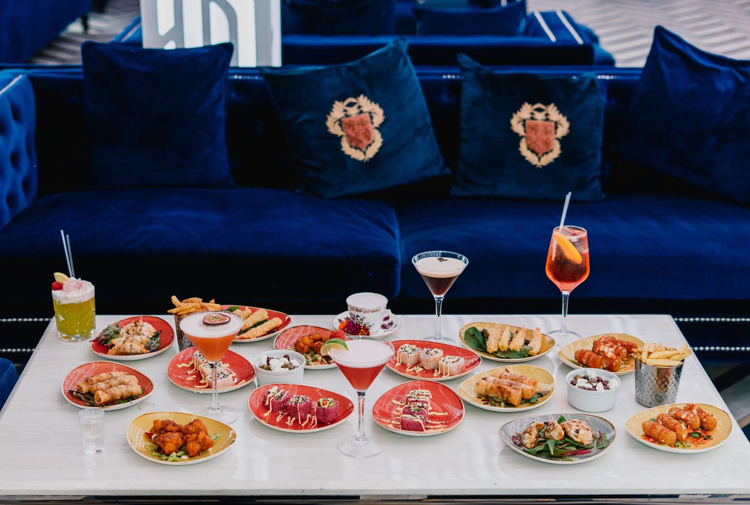A table filled with various colorful dishes and drinks, viewed from the front with a navy blue sofa and decorative pillows in the background.