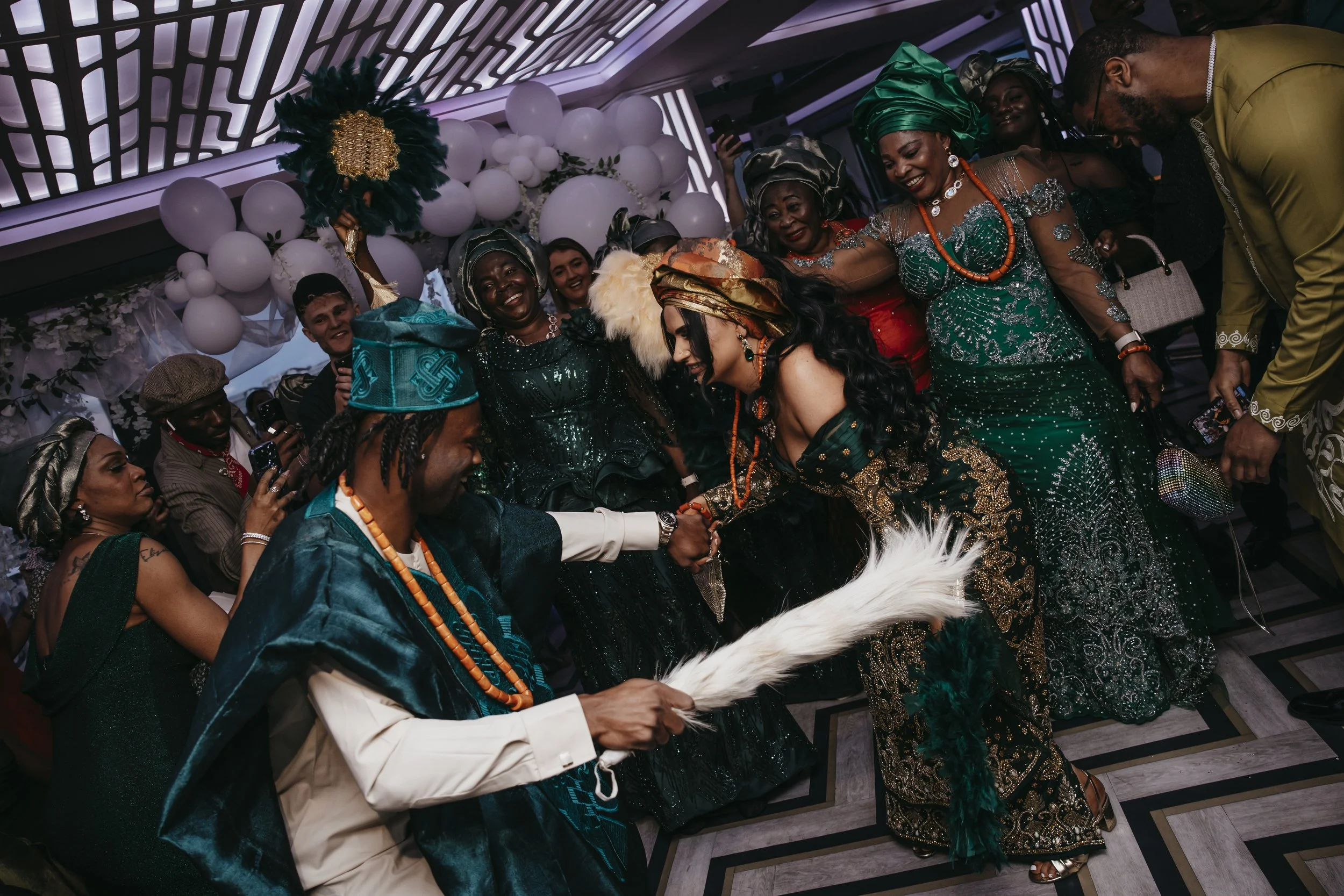 People in colorful traditional African attire celebrating at a social event, with balloons and decorative lighting in the background.