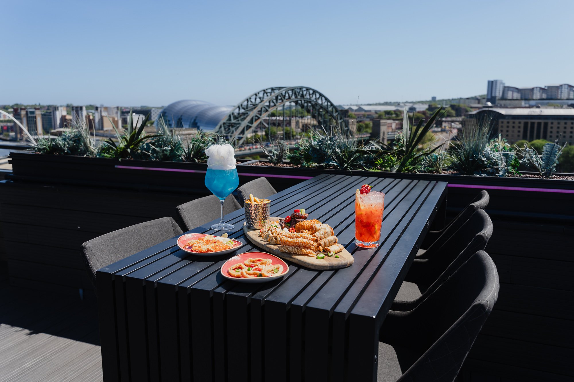Outdoor rooftop dining table with plates of pizza, fried foods, and drinks, overlooking a cityscape with a bridge and modern buildings under a clear blue sky.