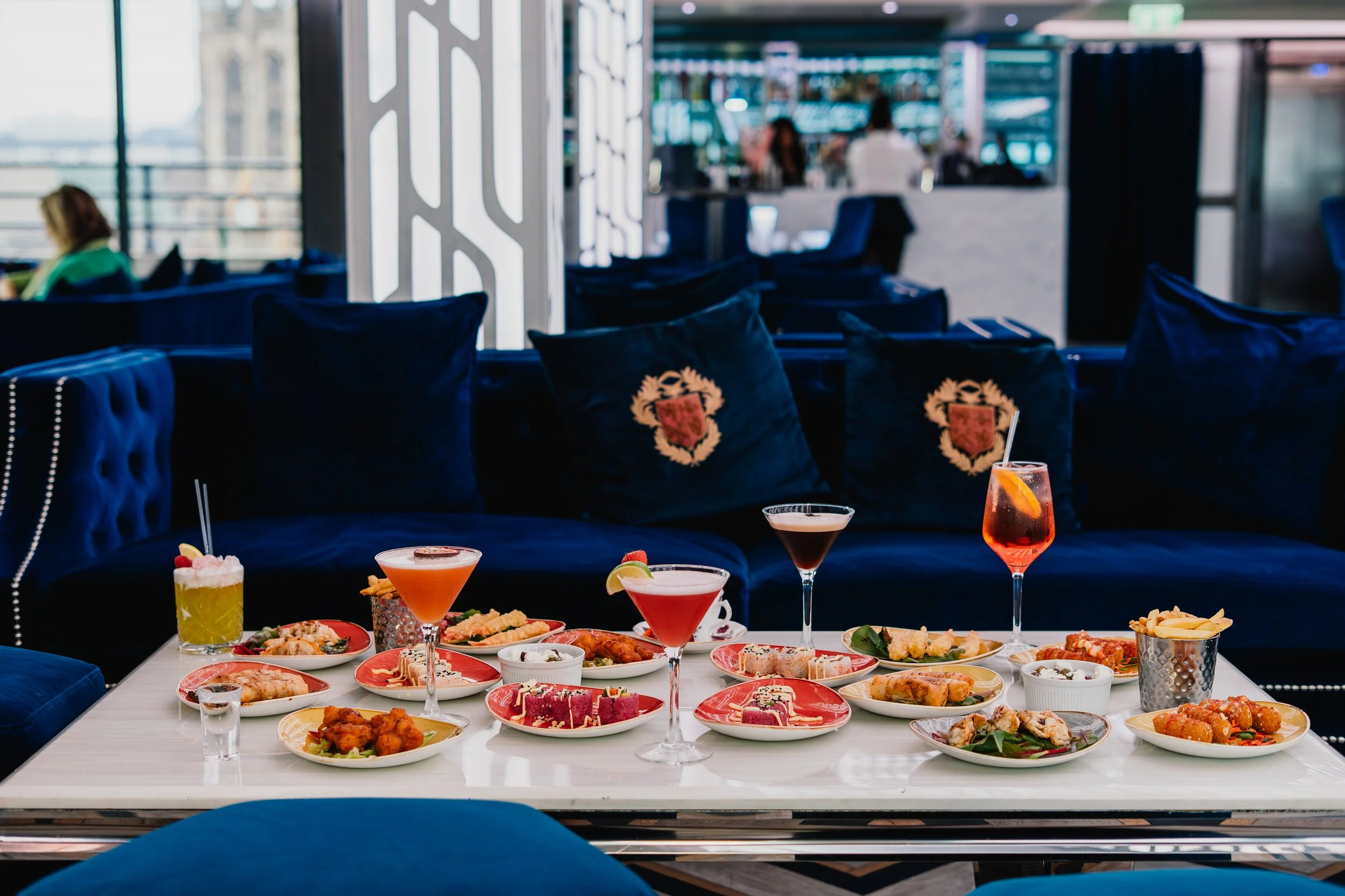 A table filled with various appetizers and colorful drinks in a bright, modern dining area with blue chairs and large windows.