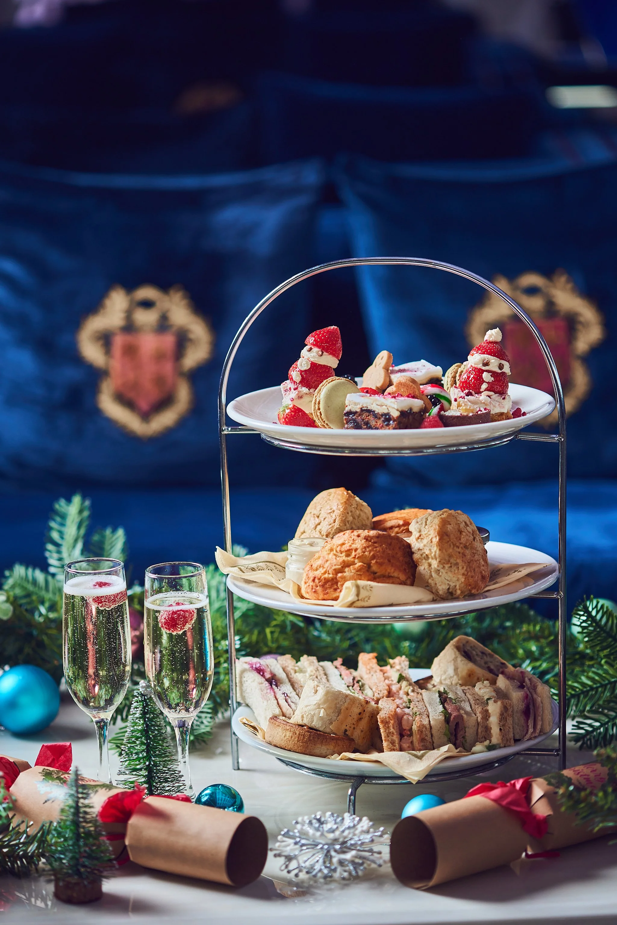 Three-tiered dessert stand with Christmas-themed treats including strawberries decorated as Santa, assorted cookies, pastries, and sandwiches. There are glasses of champagne, Christmas ornaments, gift wrap, and holiday decorations around the table.