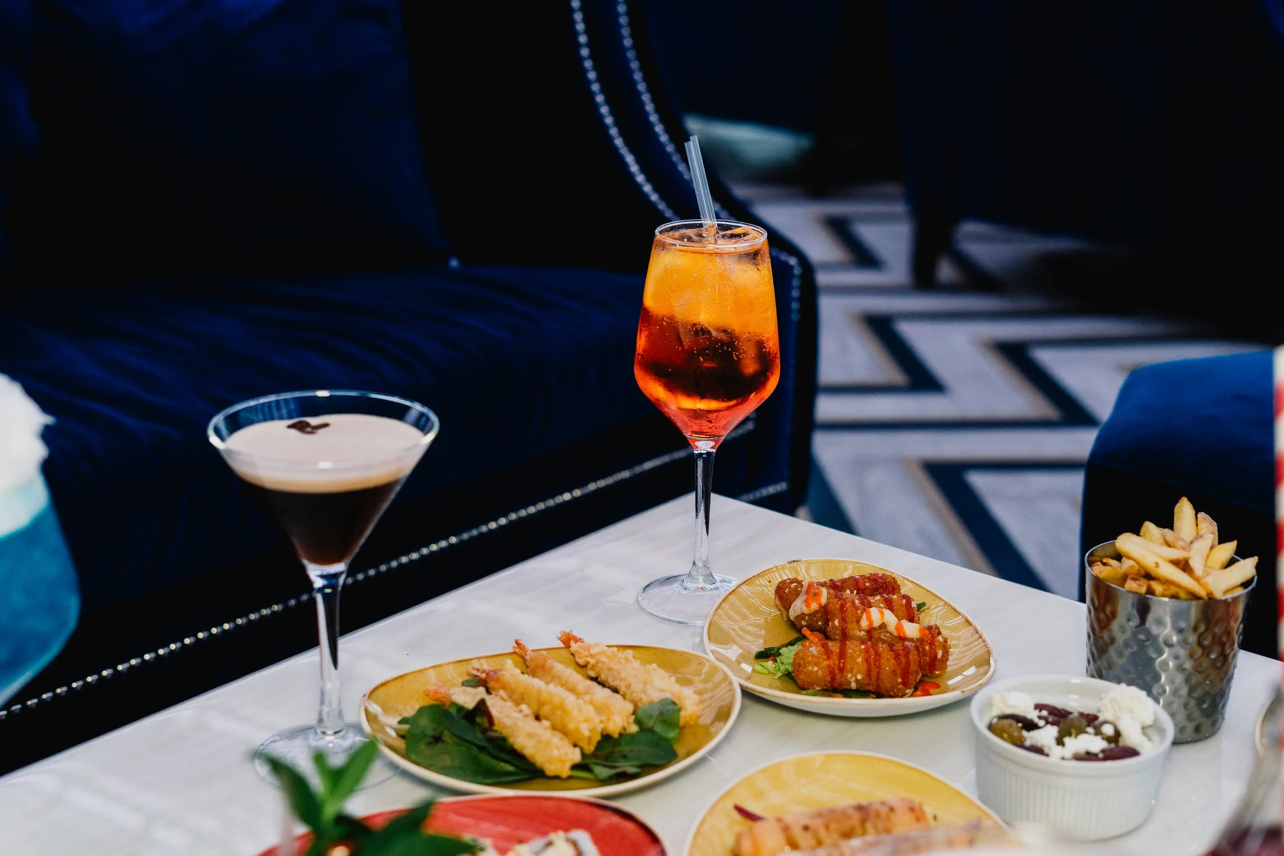 A table set with various dishes and drinks, including a glass of orange-colored beverage with a straw, a cocktail, and plates of sushi and fries, with a dark background and part of a person’s arm visible.