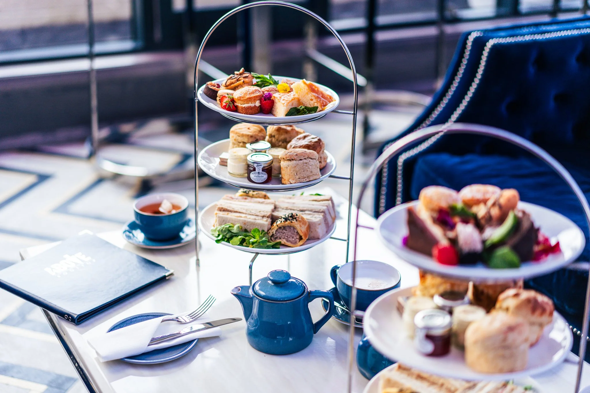 A three-tiered serving tray filled with assorted pastries, sandwiches, and fruit, set on a white table with tea cups and utensils, likely at a high tea or afternoon tea event.