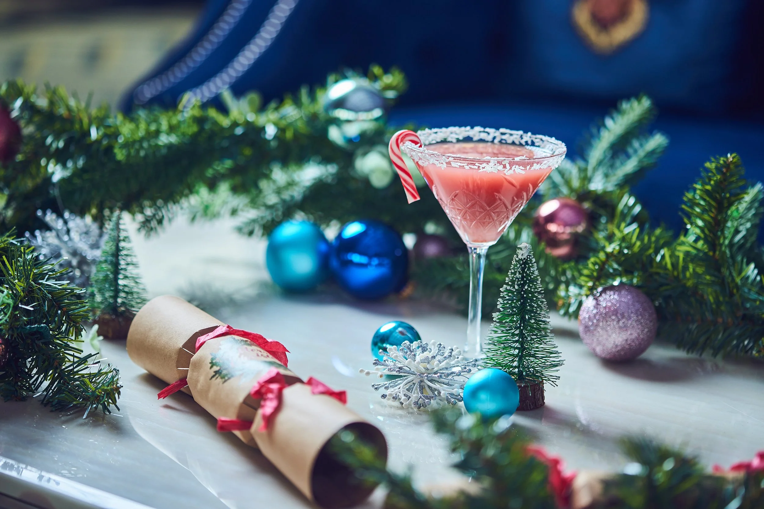 A festive holiday scene with a pink cocktail topped with a peppermint candy cane, surrounded by Christmas decorations including green pine branches, colorful ornaments, a silver snowflake, and Christmas crackers.