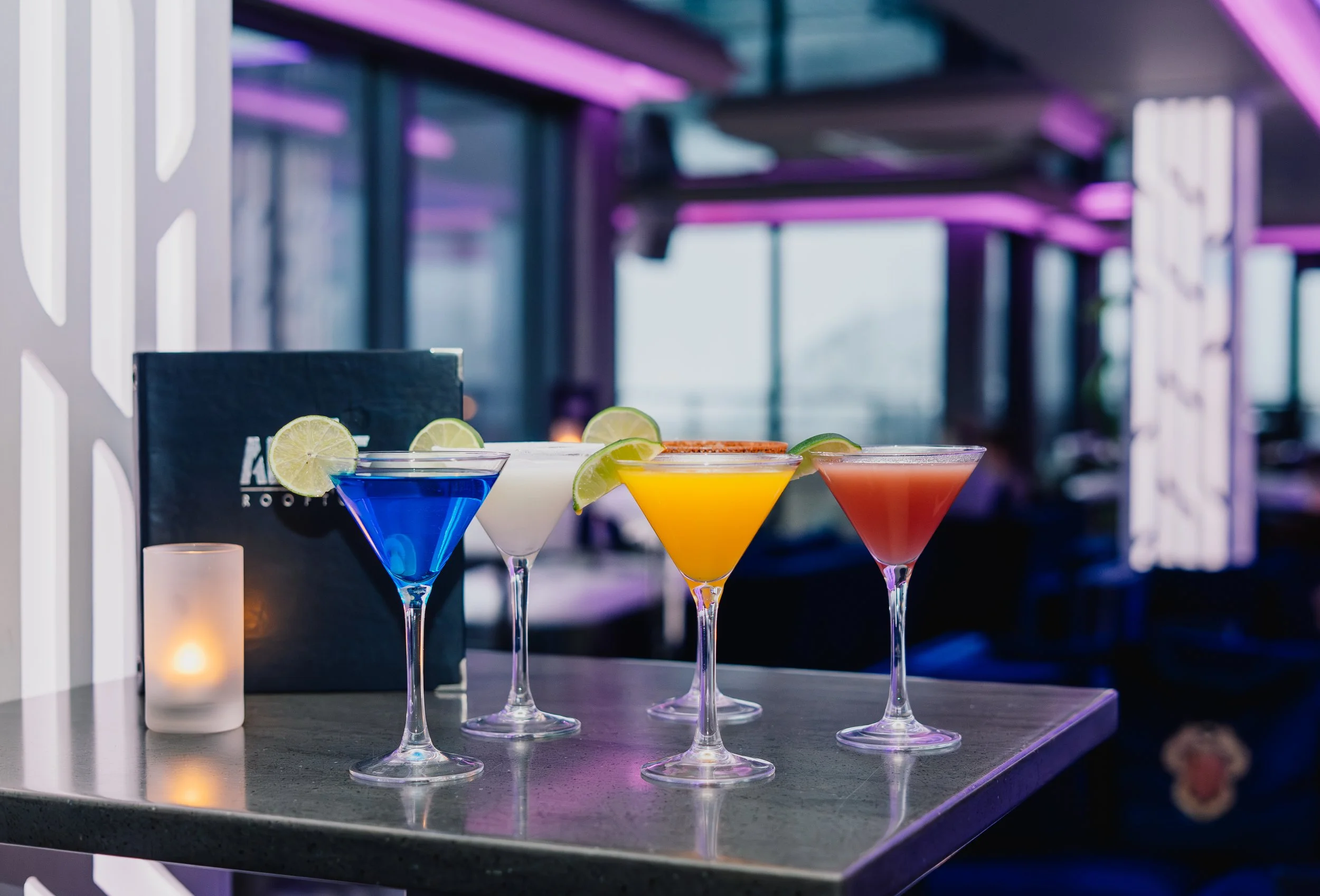 Four colorful cocktails in martini glasses with lime garnishes on a bar counter, with a blurred purple-lit background.