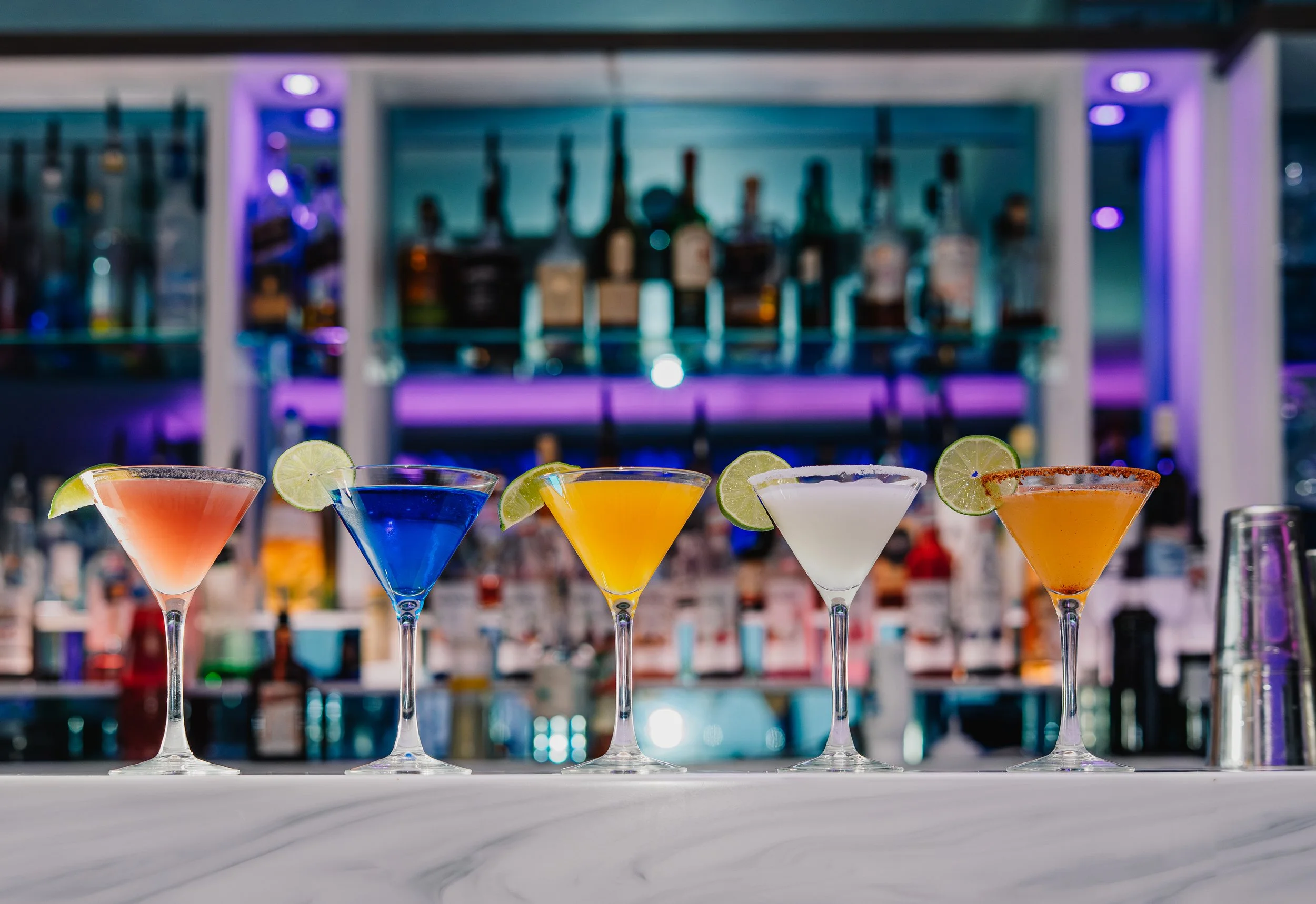 A row of colorful cocktails garnished with lime slices, set on a bar counter in front of a neon-lit bar shelf with various bottles.