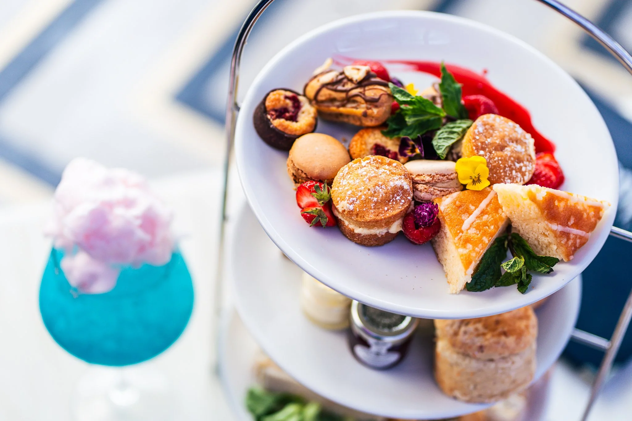 Tiered serving tray with assorted mini desserts including cookies, cake slices, and pastries, garnished with mint leaves and edible flowers, with a blurred pink cupcake in a blue wrapper visible in the background.