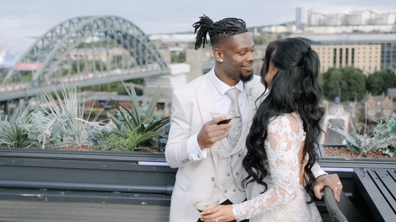 A man and woman in wedding attire sharing a romantic moment with drinks on a rooftop with city skyline and a bridge in the background.