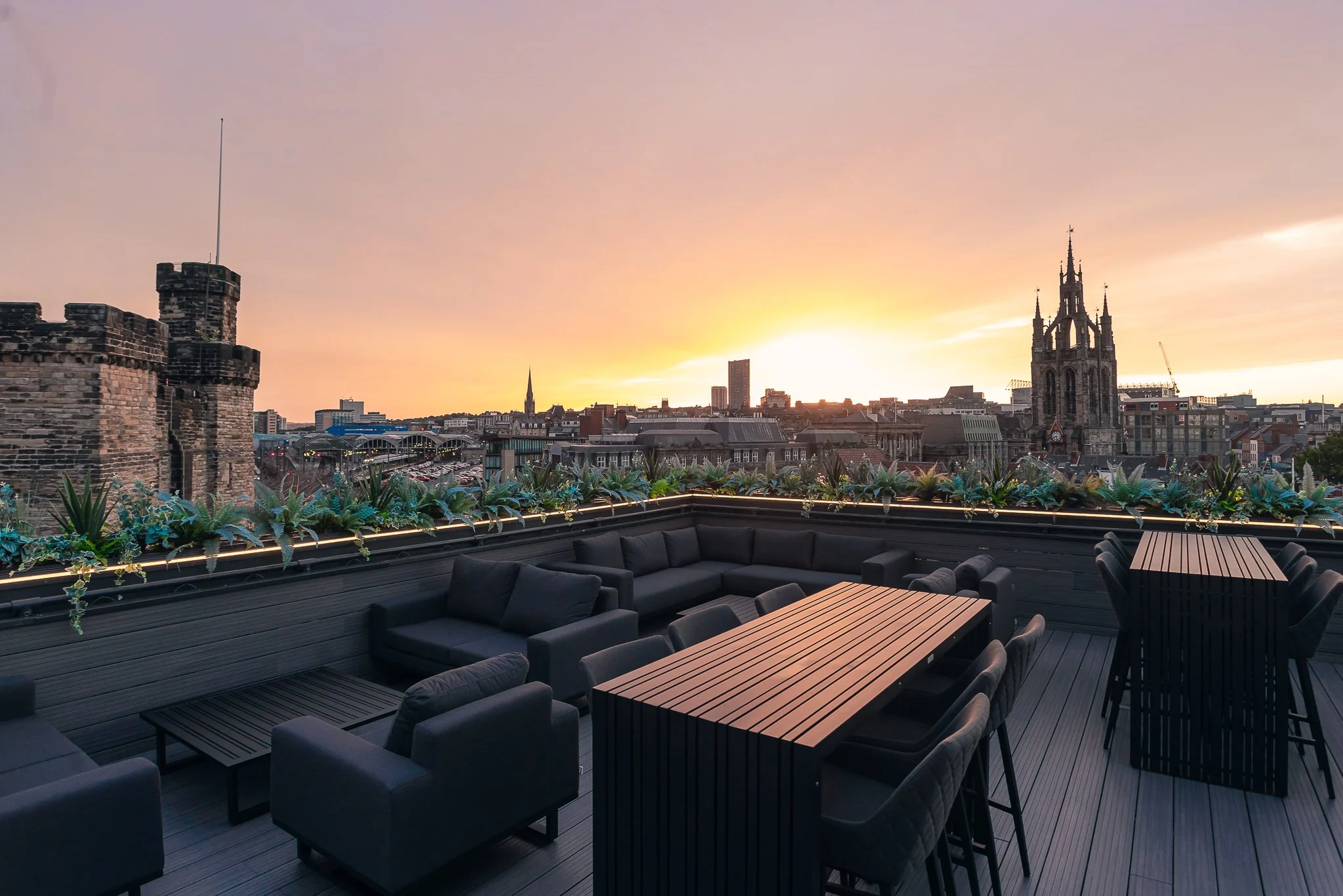 Rooftop lounge with black outdoor seating, potted plants, and a view of a city skyline at sunset, including historic and modern buildings.