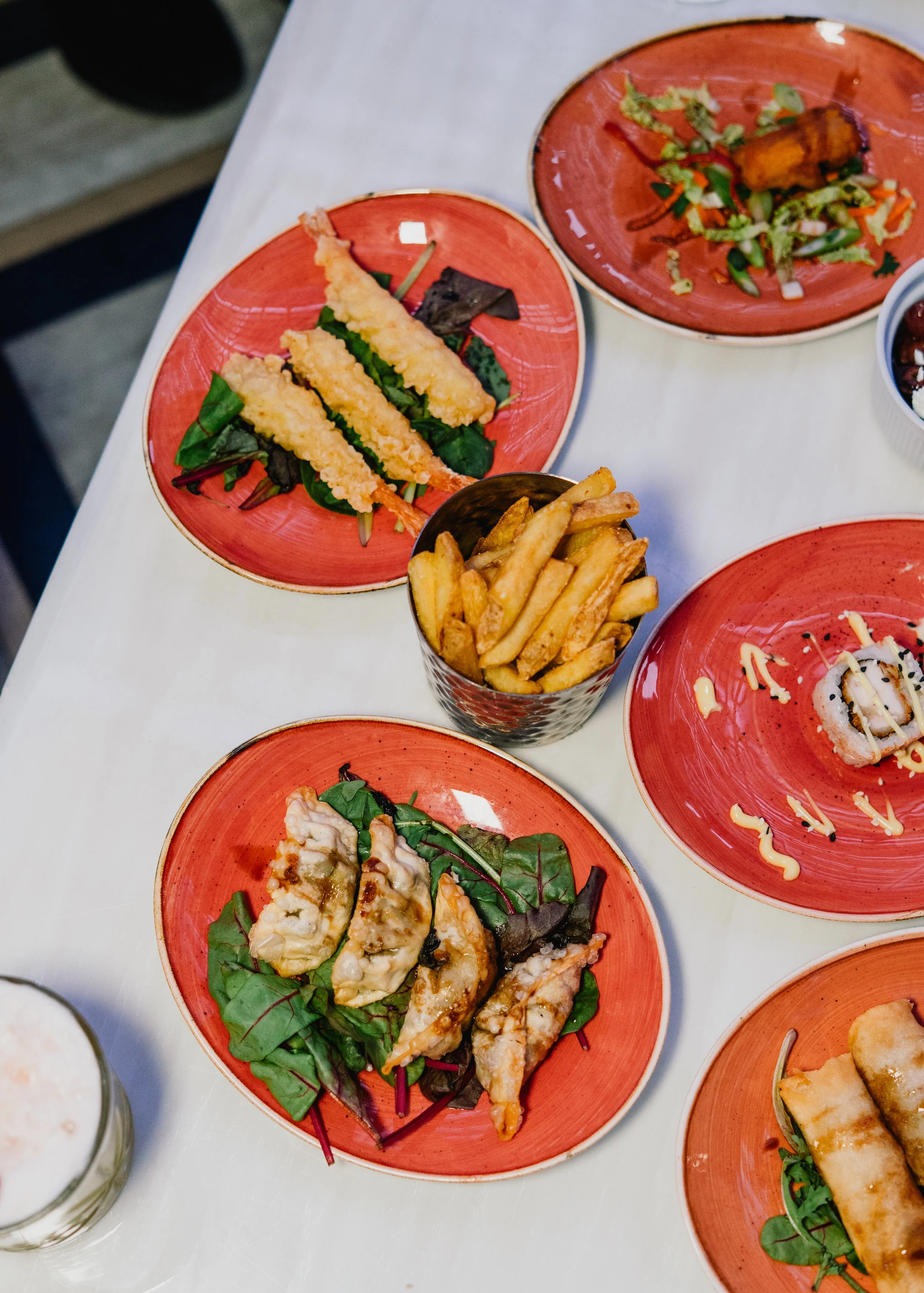 Various Asian dishes served on red plates on a white table, including fried tempura with dipping sauce, a salad with chopped vegetables, and sushi rolls with drizzle sauce.