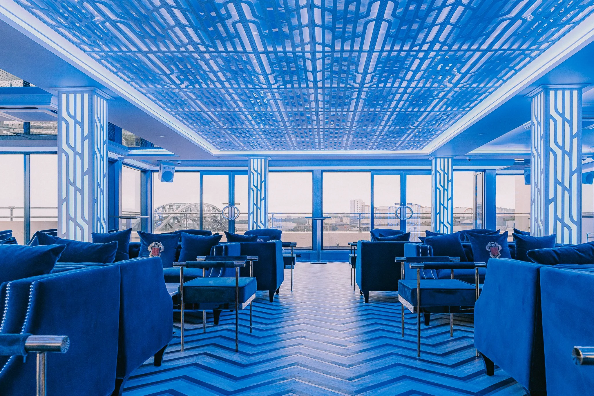 Modern interior with blue velvet chairs, large windows, and a geometric ceiling design, in a space overlooking a cityscape.