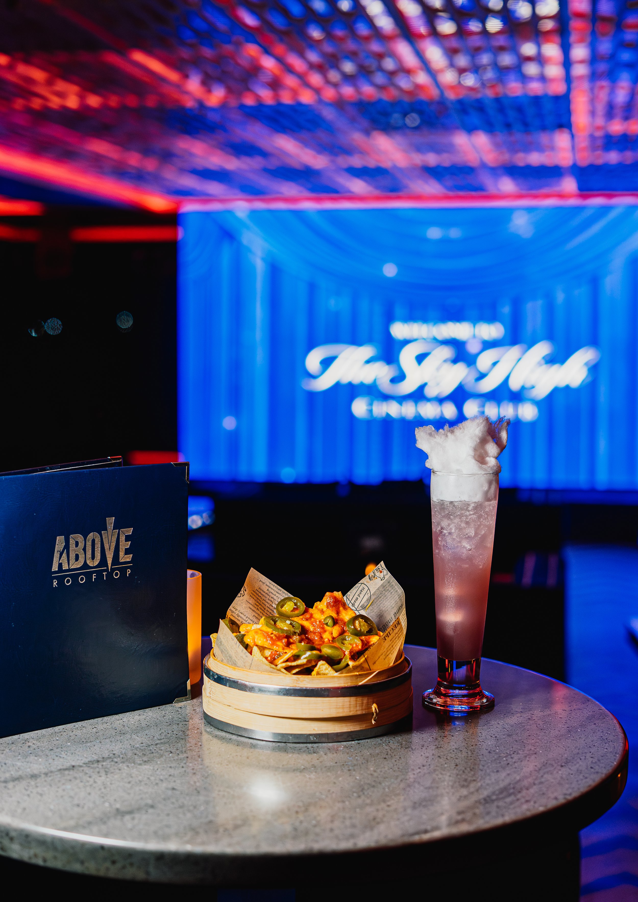 A table with a pizza topped with jalapenos, a cold beverage with ice, and a menu, in a dimly lit, neon-lit bar or nightclub.