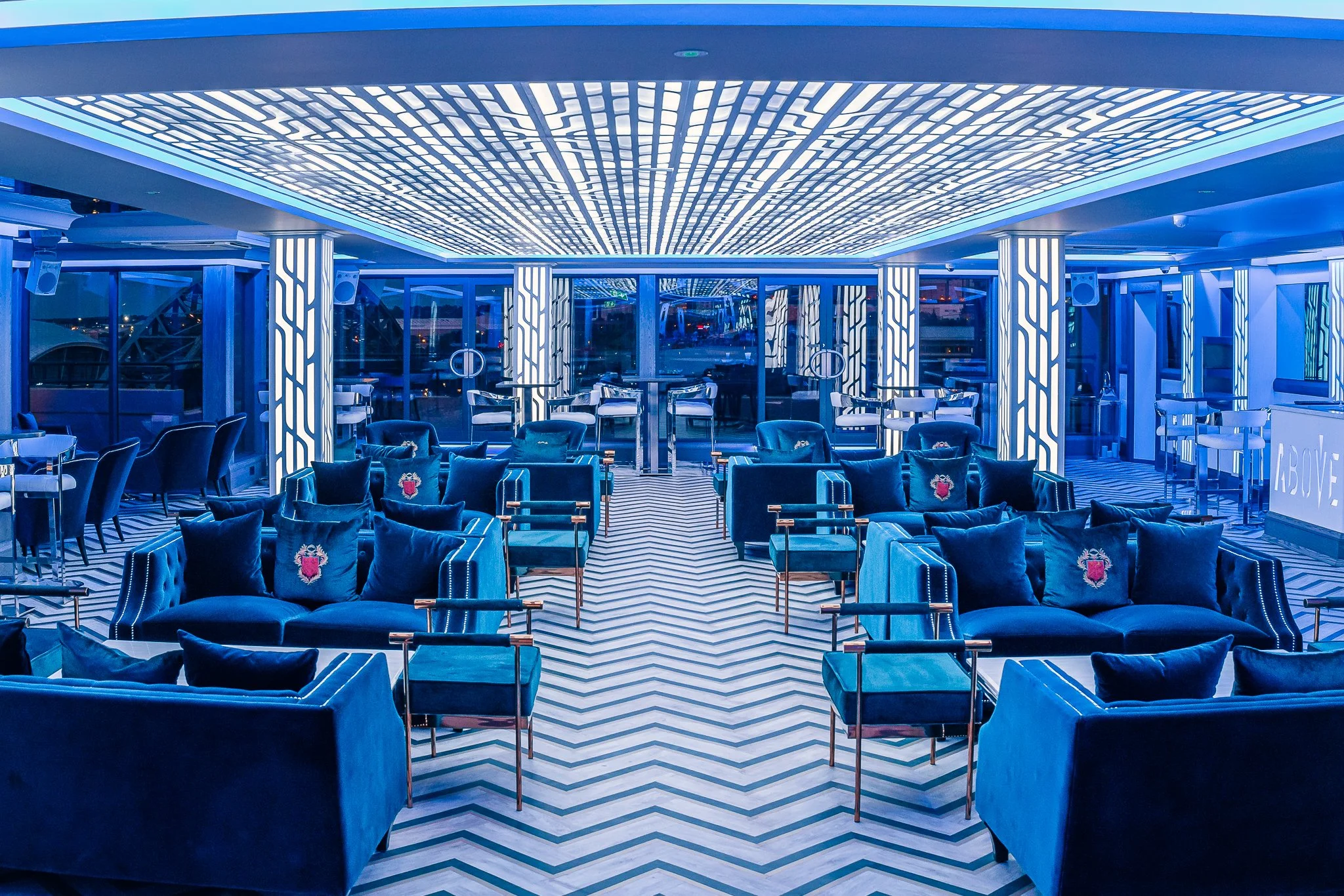 Interior of a modern, well-lit lounge area with blue velvet sofas, chairs, and tables, illuminated with blue and white LED lighting, featuring a geometric chevron-patterned carpet.
