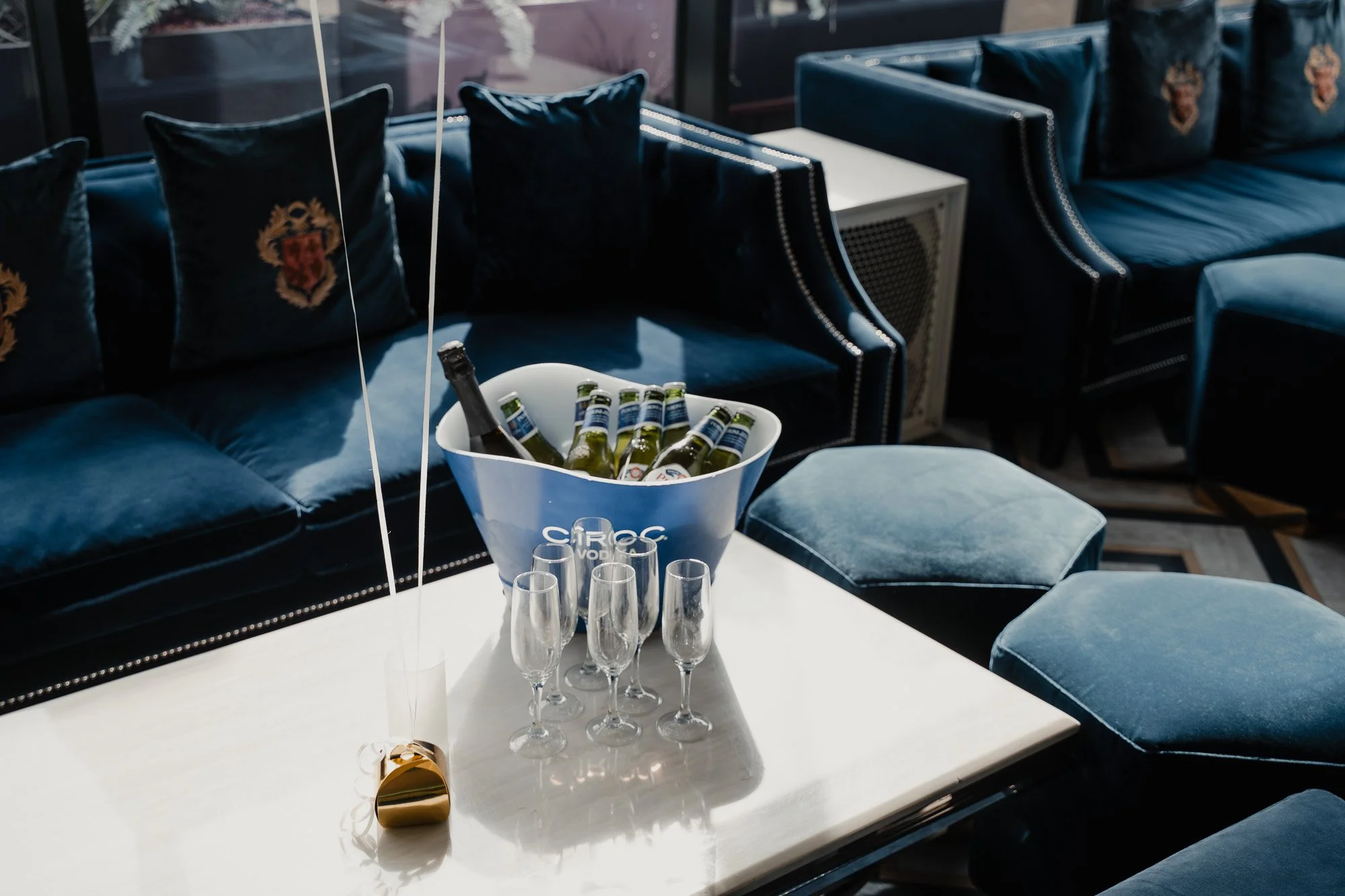 A white table with empty champagne flutes and a bucket of bottles, set in a lounge with blue velvet chairs and decorative pillows with crest emblems.