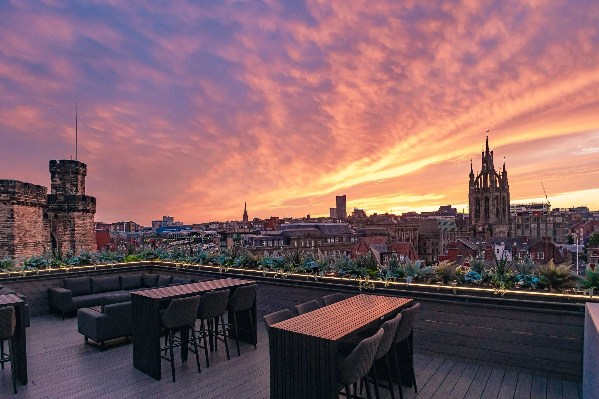 Rooftop patio with outdoor seating overlooking city skyline at sunset, featuring historic church and modern buildings, with pink and orange clouds in the sky.