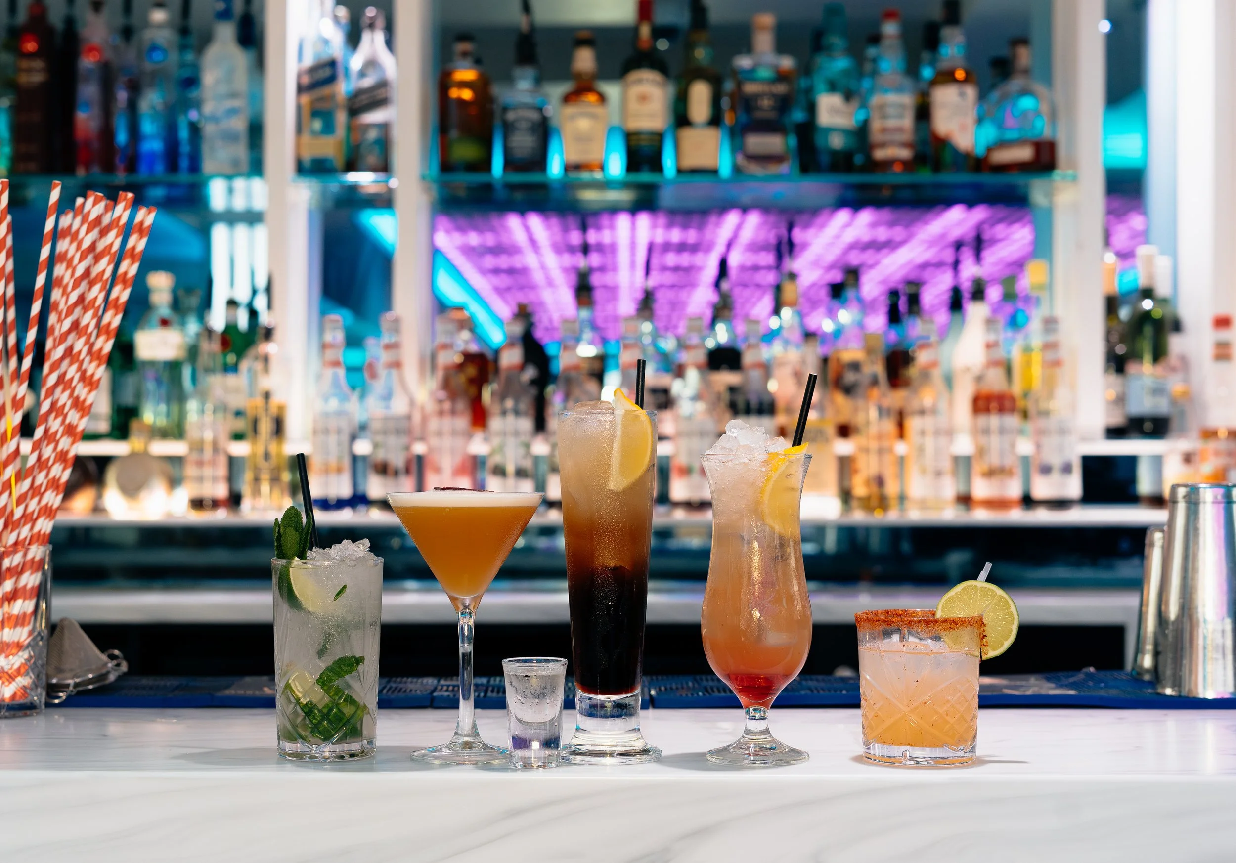 A variety of colorful cocktails on a bar counter with blurred liquor bottles and neon lighting in the background.