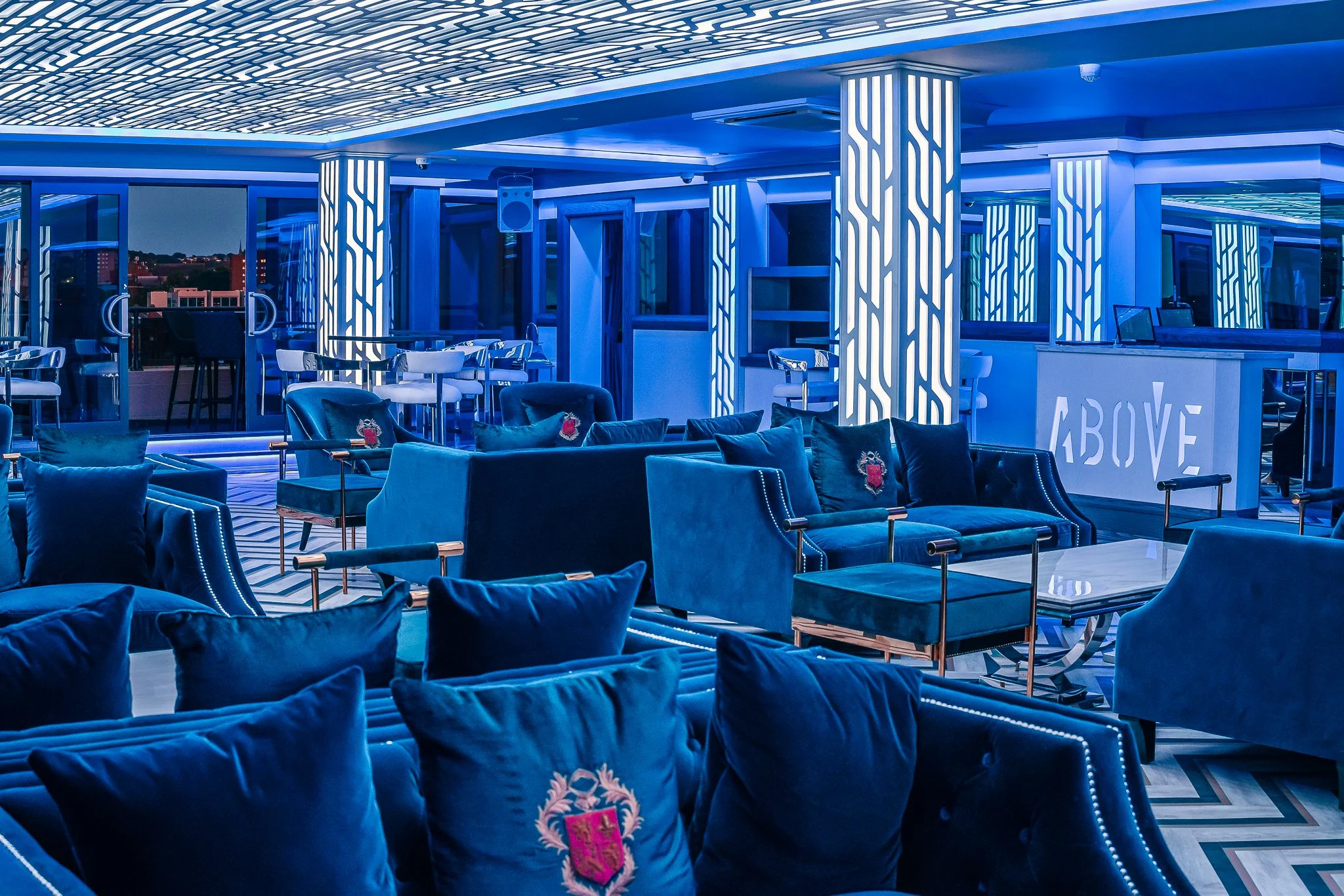 A modern, stylish lounge area with blue velvet chairs and pillows, illuminated by blue lighting, featuring a bar area with a sign that says 'ABOVE', and large windows showing city buildings at night.