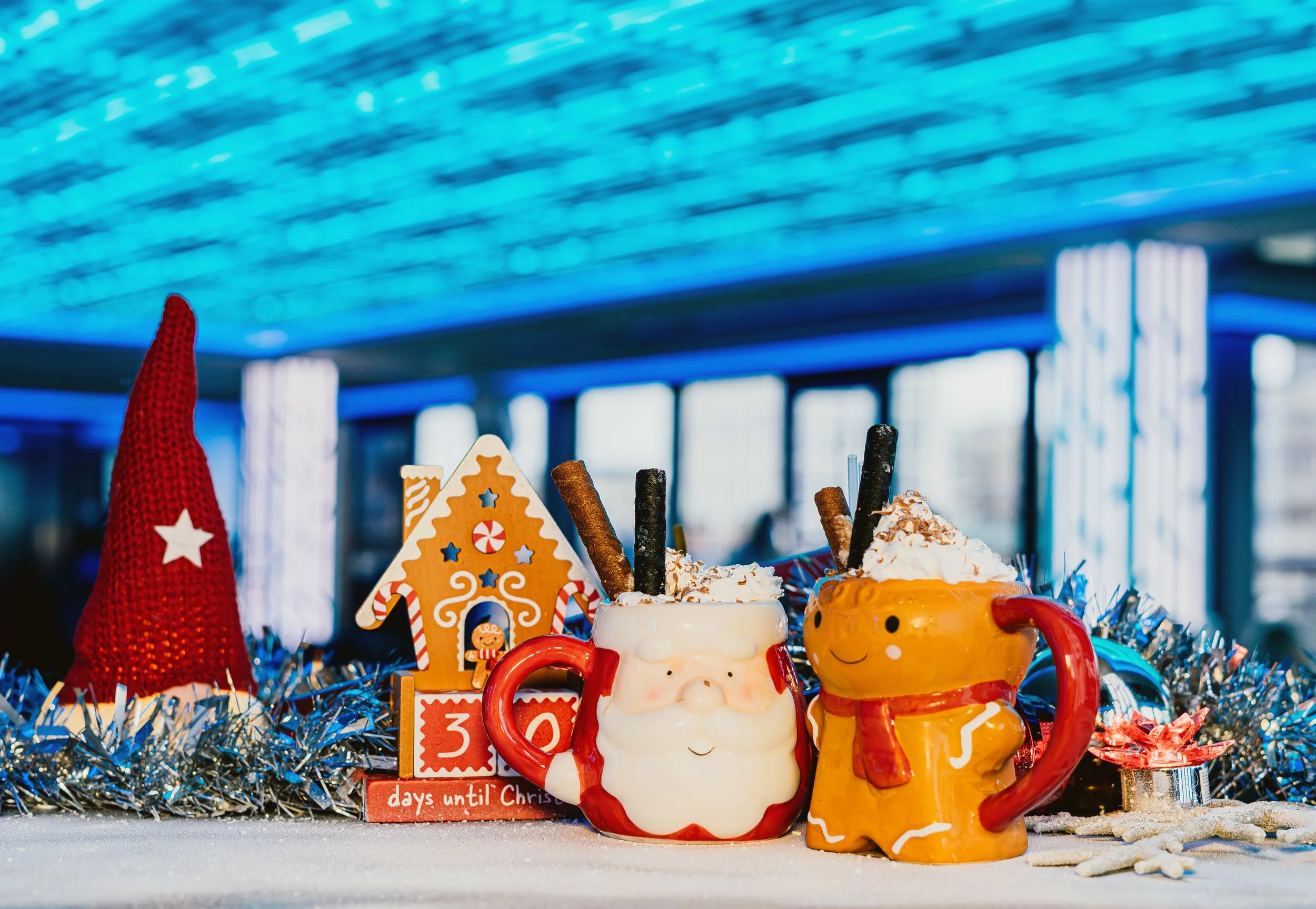 Colorful holiday mugs and gingerbread cookies on a festive table with a snow-like surface and decorative tinsel, in front of a blue-lit ceiling.