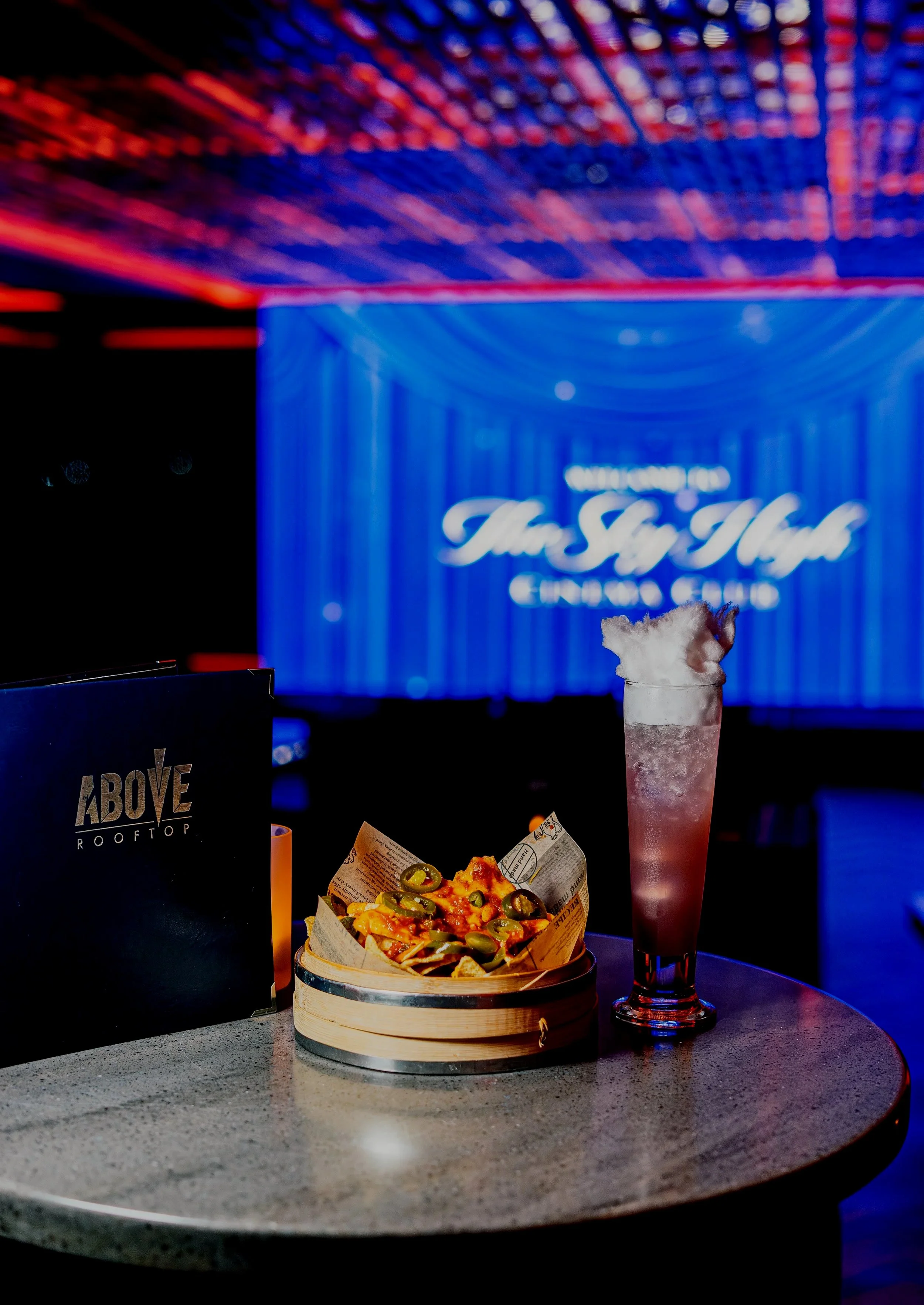 A pizza topped with jalapeño peppers, a drink with ice, and a black menu on a round table in a nightclub or bar with blue and red lighting.
