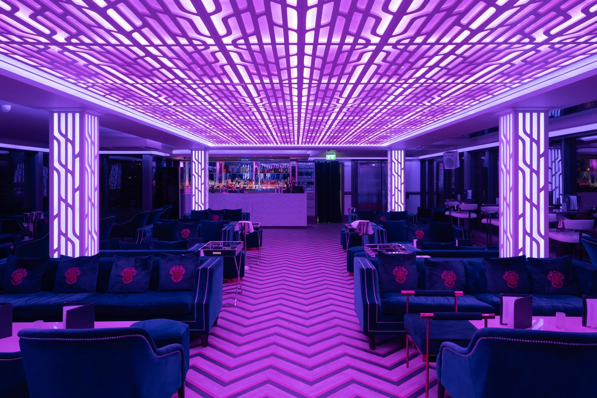 A nightclub interior with purple neon lights, patterned ceiling, and seating area with velvet chairs and small tables.