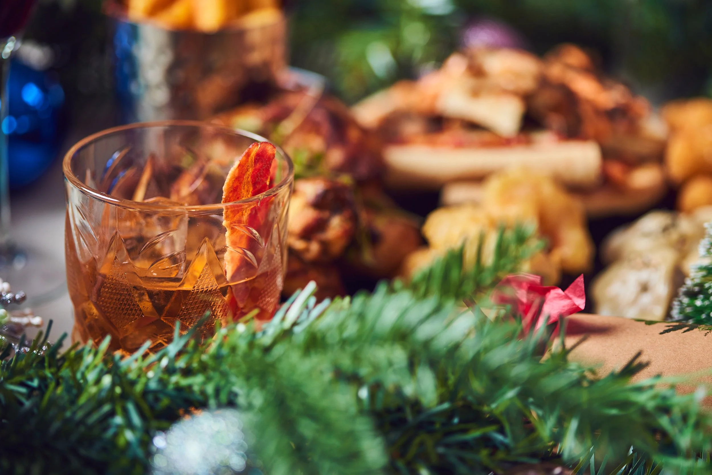 A glass of brown liquid with a slice of dried apple in it, placed on a decorated surface with blurred snacks and greenery in the background.
