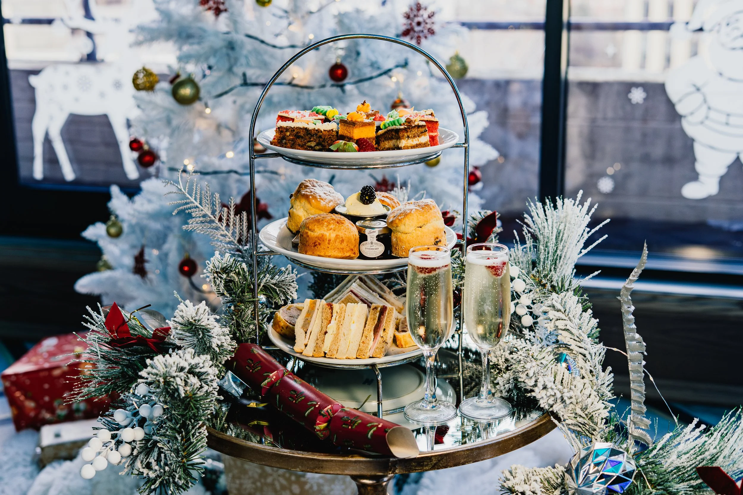 A festive three-tiered serving stand with assorted desserts on a decorated table in front of a snowy Christmas tree. The top tier has slices of cake with colorful icing, the middle tier holds scones, and the bottom tier features finger sandwiches. Th