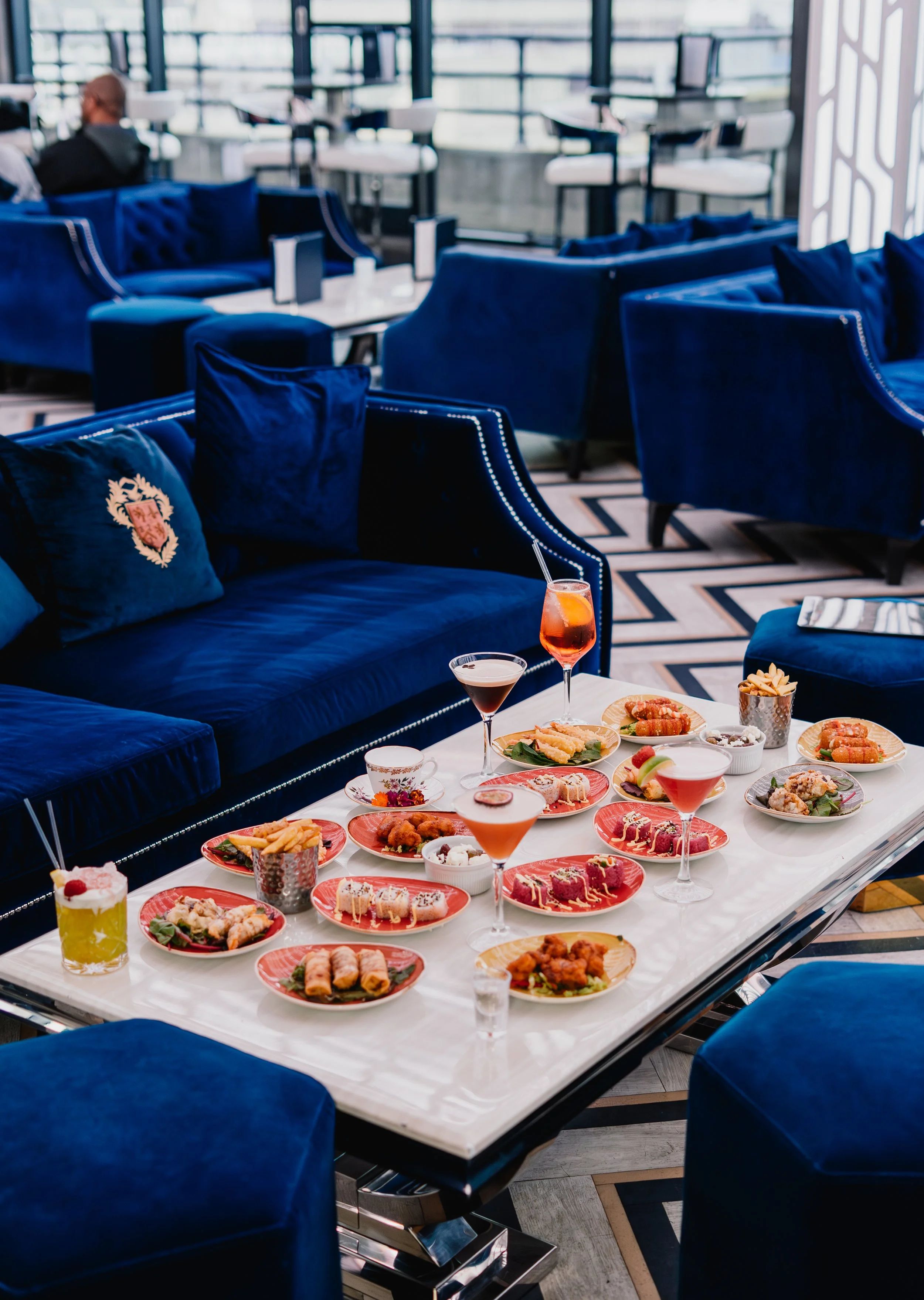 A white table filled with various appetizers, desserts, and drinks in a modern lounge with blue velvet chairs and large windows.