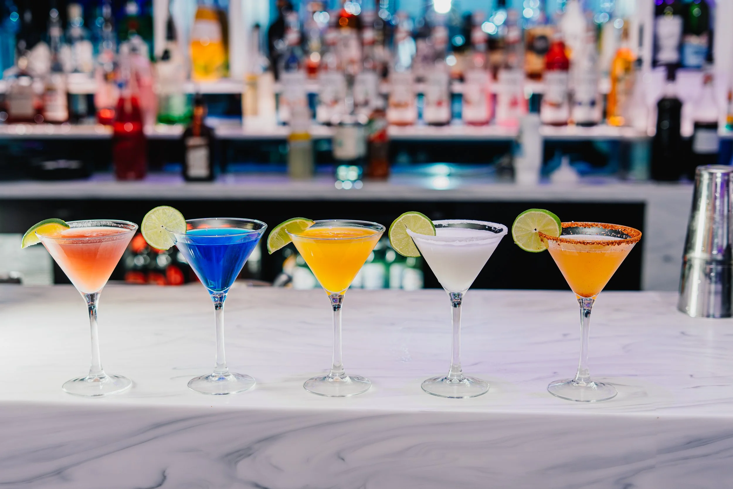 Five colorful cocktails with lime wedges on a bar counter with a blurred bar background.