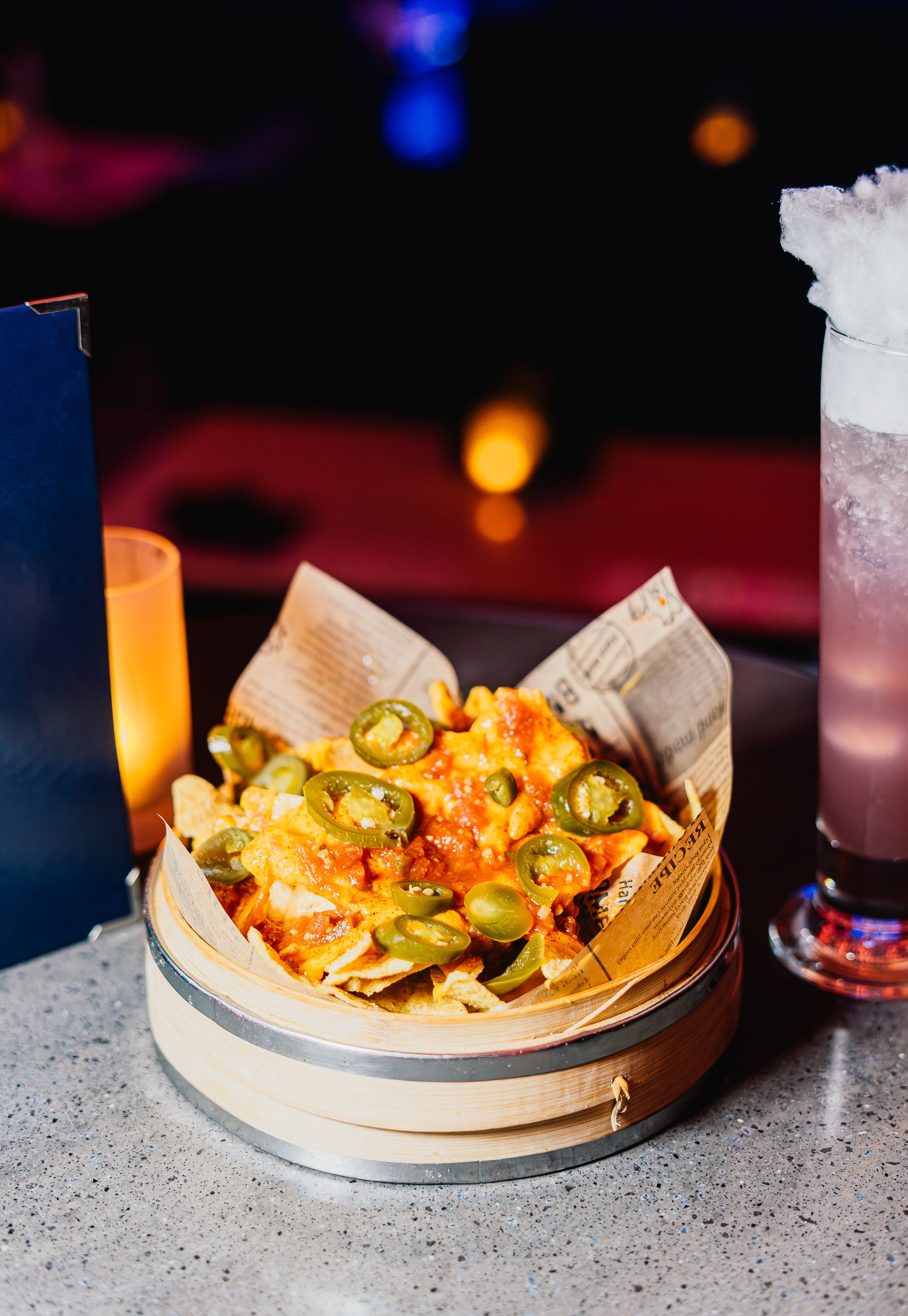 A slice of pizza with green peppers on top, served in a paper-lined basket on a round wooden tray, with a lit candle and a glass of pink drink nearby.