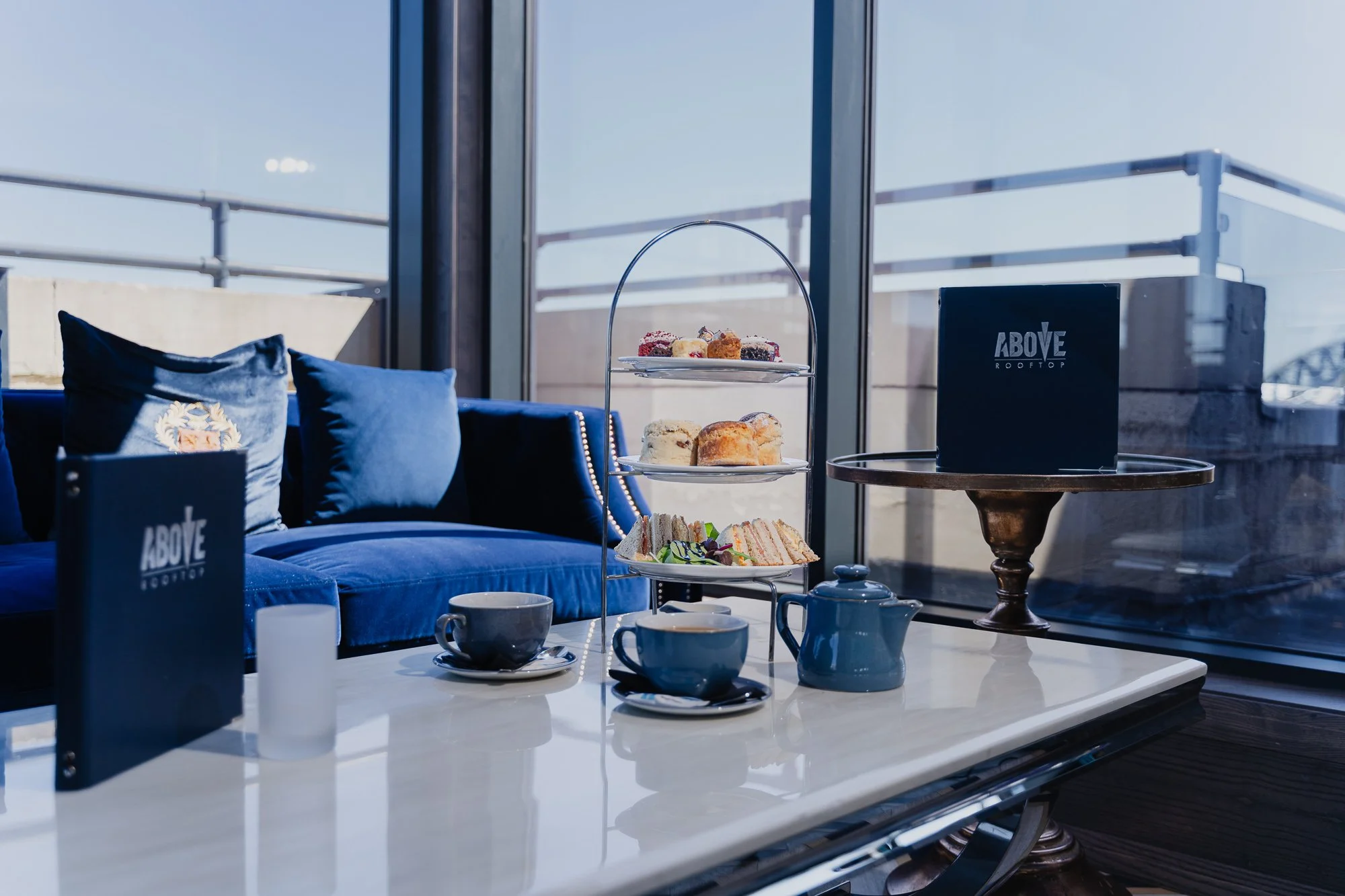A table with cups, a three-tiered tray of assorted desserts, sandwiches, and a black box labeled 'Above Rooftop' in an indoor lounge area with large windows, blue pillows, and outdoor structures visible in the background.