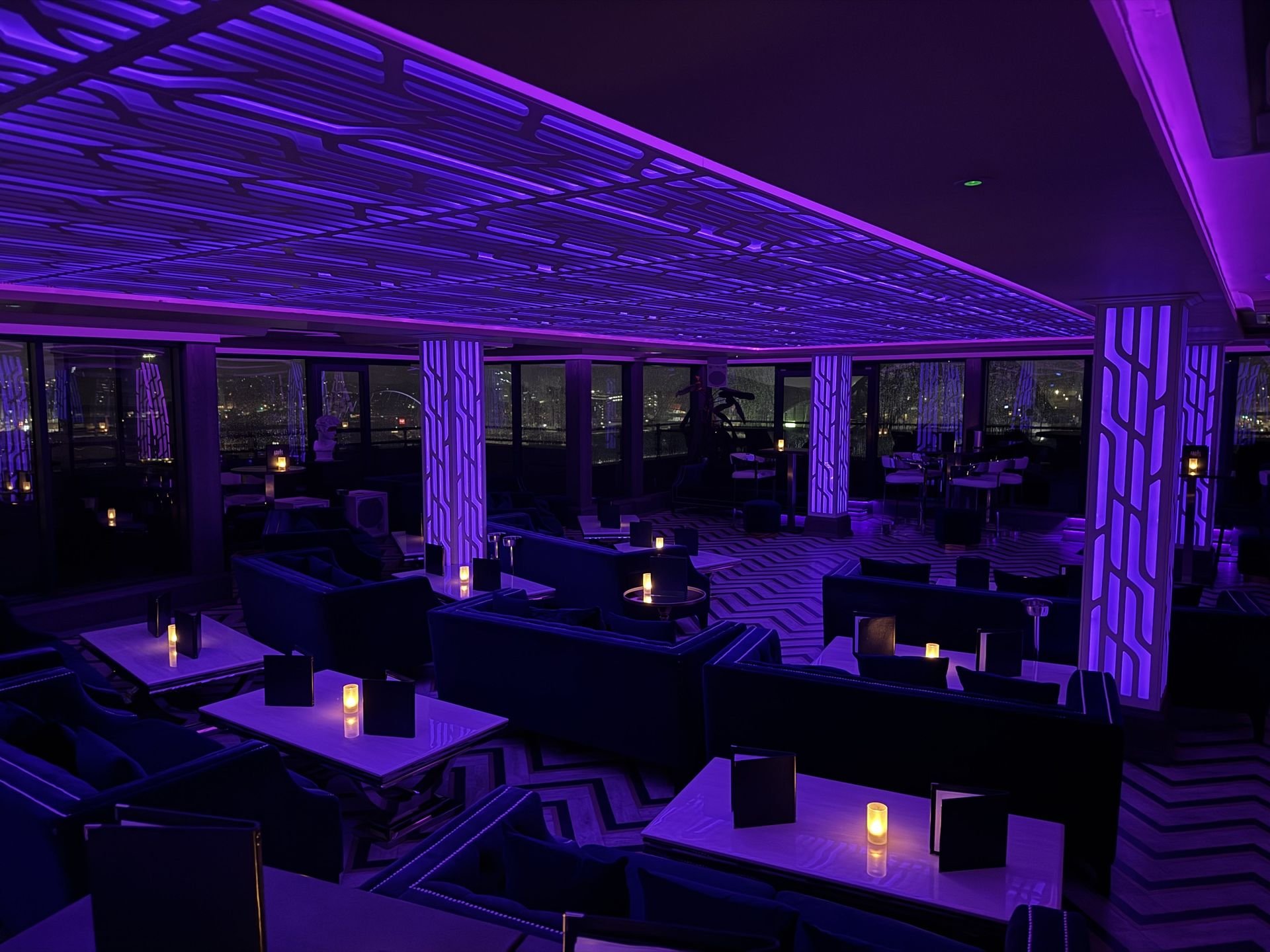 Inside a dimly lit lounge with purple neon lighting, modern seating, tables with candles, and large windows showing city lights at night.