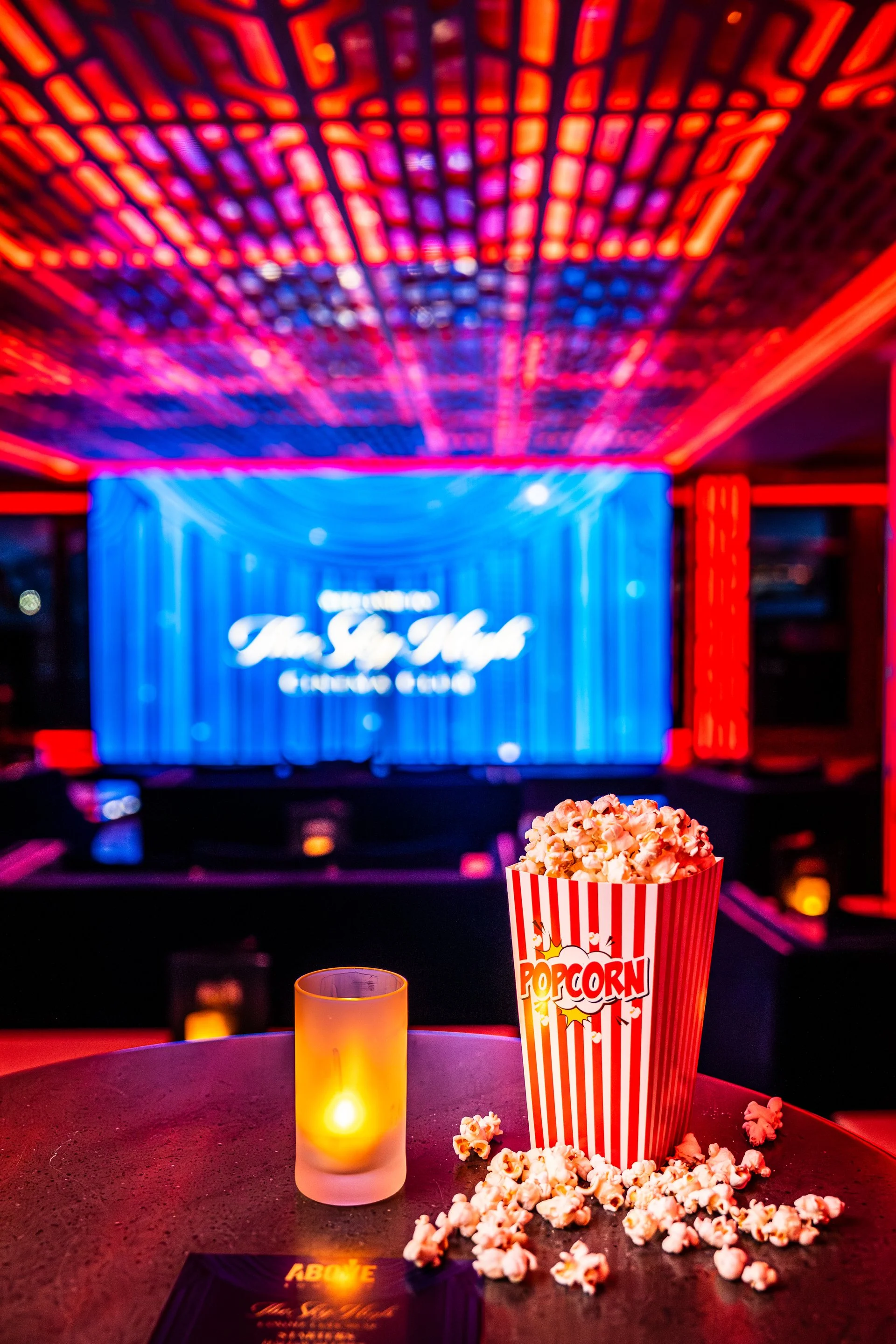 A container of popcorn on a table at a movie theater, with a neon-lit ceiling and a blue screen in the background.