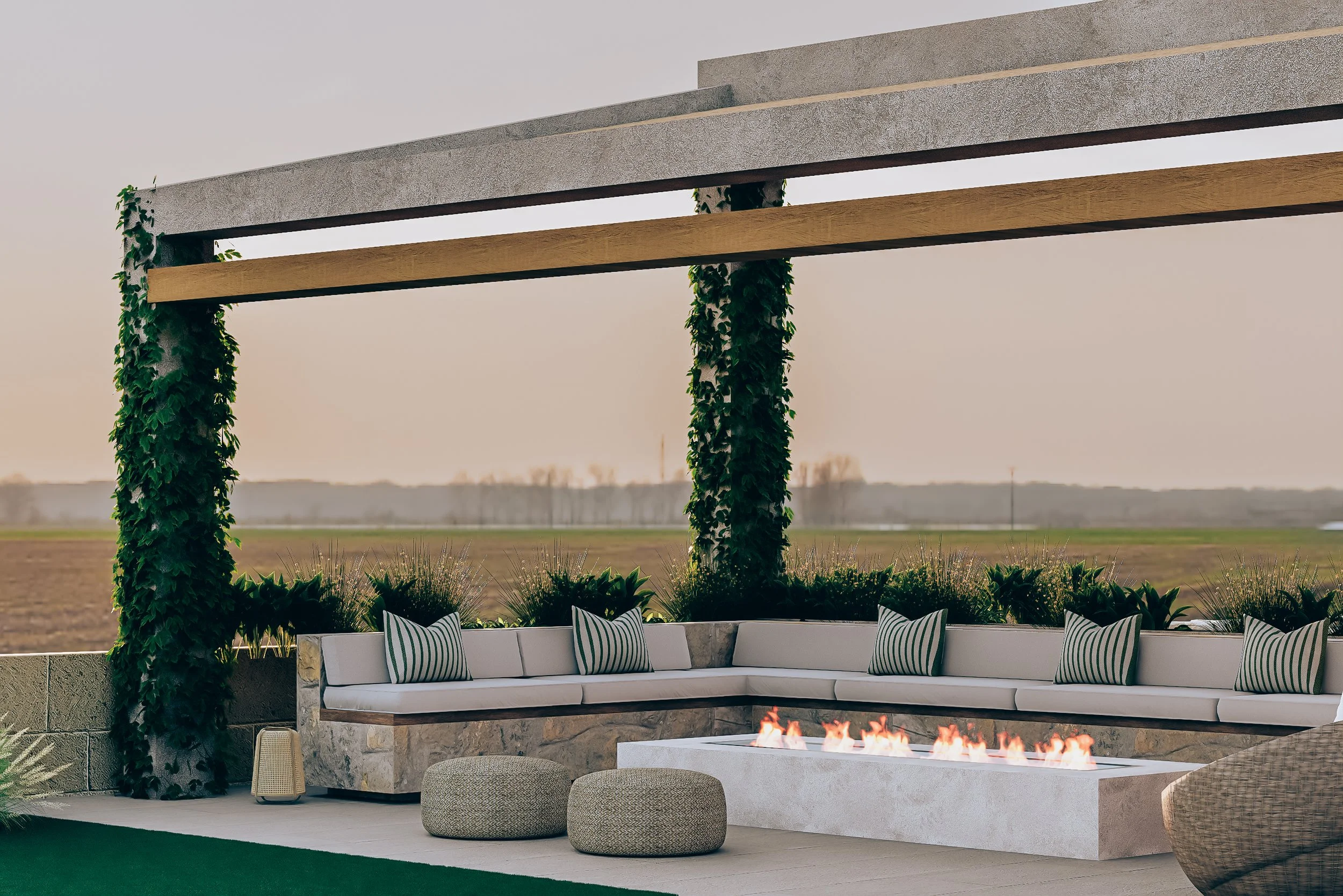 Modern outdoor patio with a built-in fireplace, white seating with striped cushions, and greenery against a field landscape.