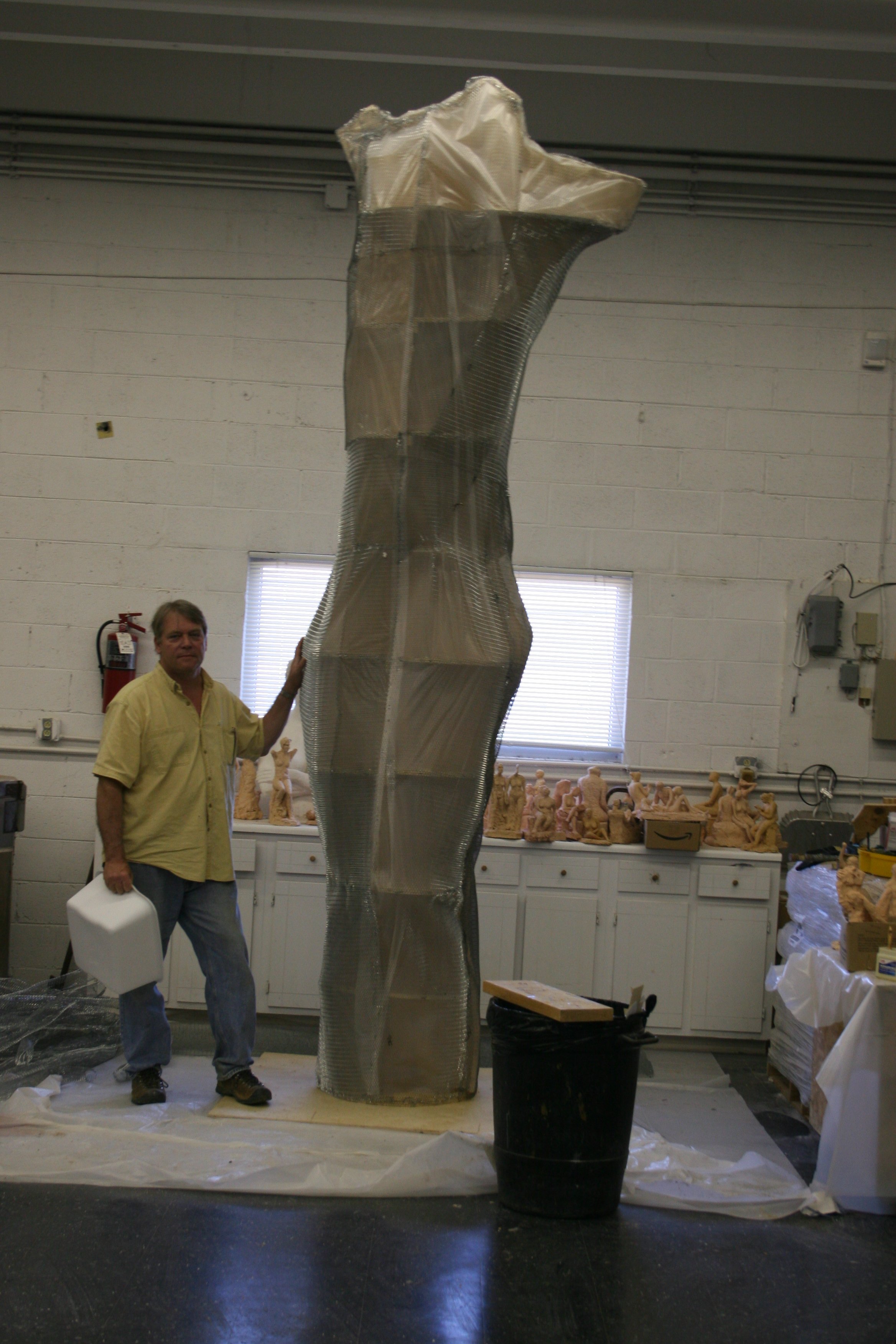 A man in a yellow shirt standing next to a tall, abstract metallic sculpture in an indoor space with white walls and a garage-style window. The sculpture is being held by the artist Alan LeQuire.