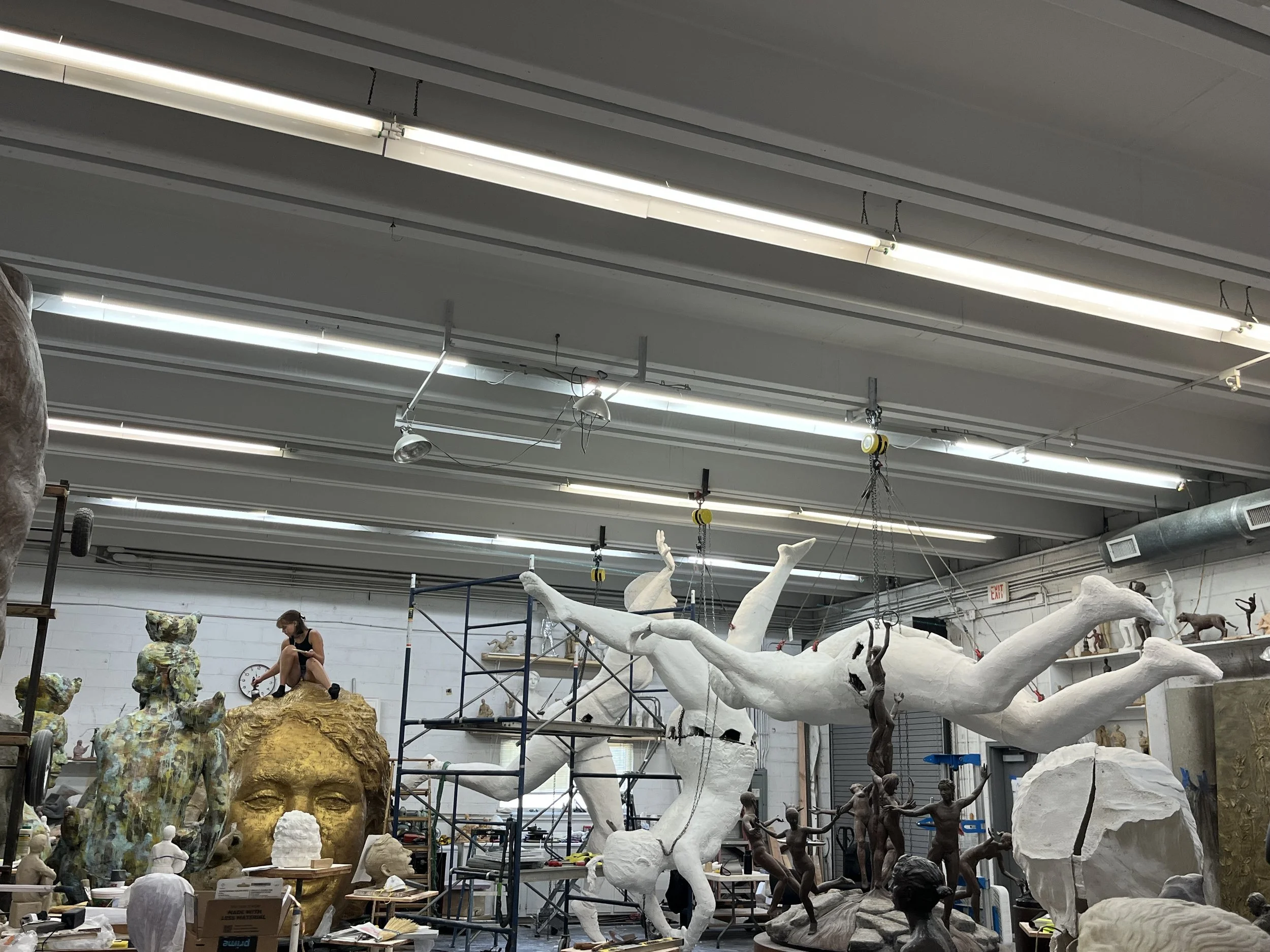 A spacious workshop with various sculptures in different stages of completion, including a large white reclining figure of a person and a gold-colored head sculpture, with tools and scaffolding around.