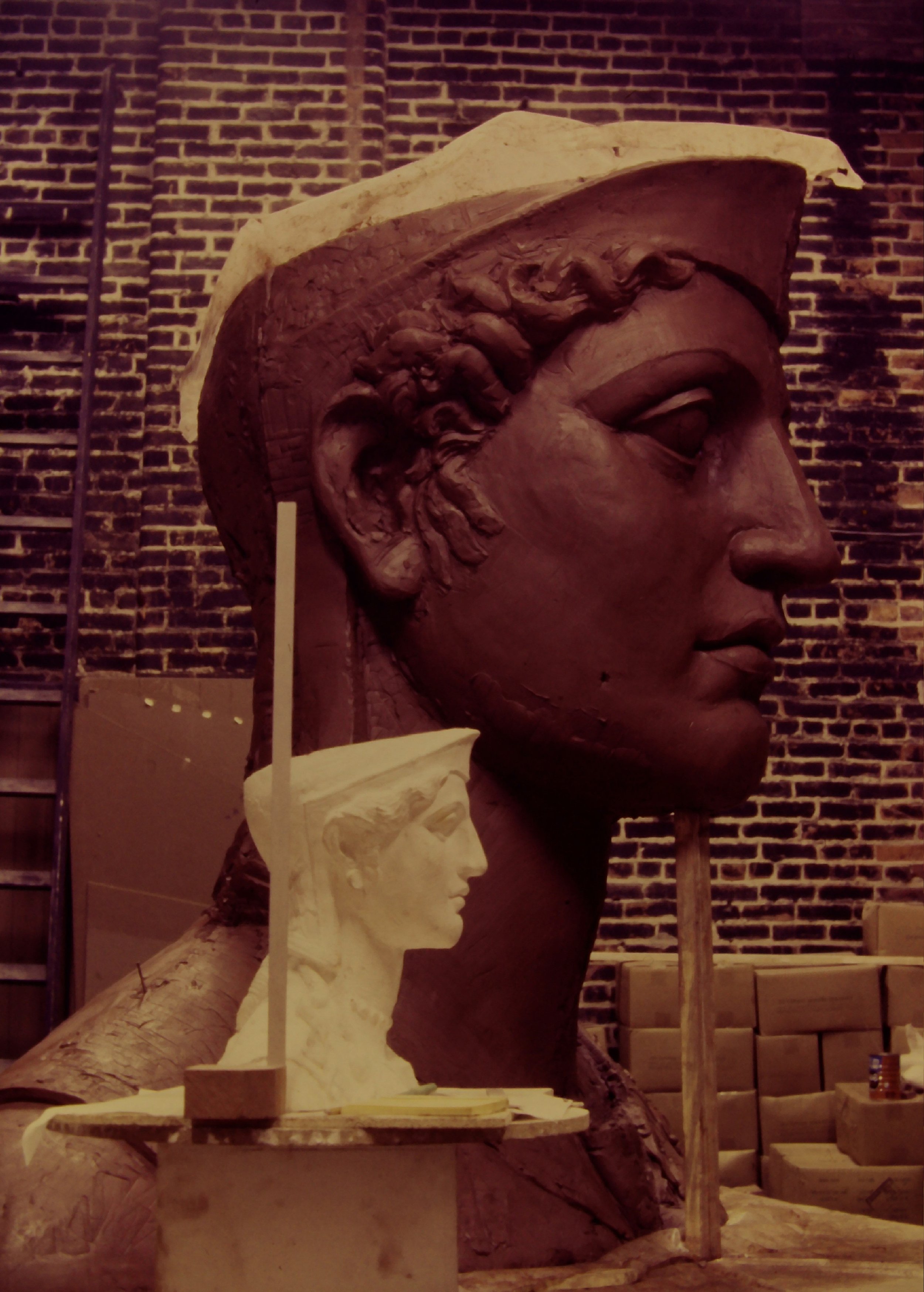 A large stone sculpture of a person's head in profile views, with detailed facial features and a textured hairstyle. A white plaster bust of a woman's head is attached to the smaller side of the sculpture. Athena Parthenos in Parthenon Museum 