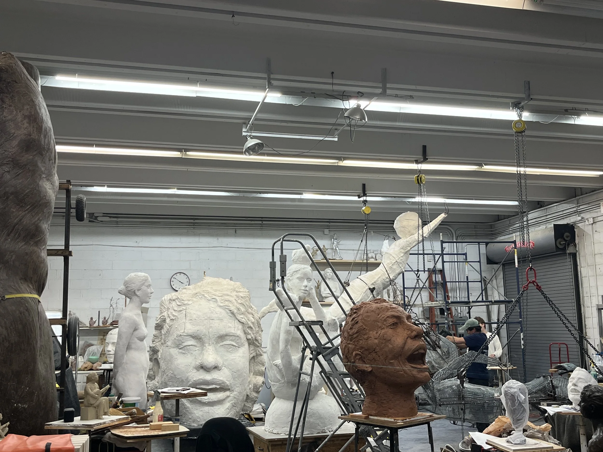 Inside a sculpture studio, various unfinished statues and busts are visible, with some artists working on sculptures. Overhead fluorescent lighting illuminates the workspace.