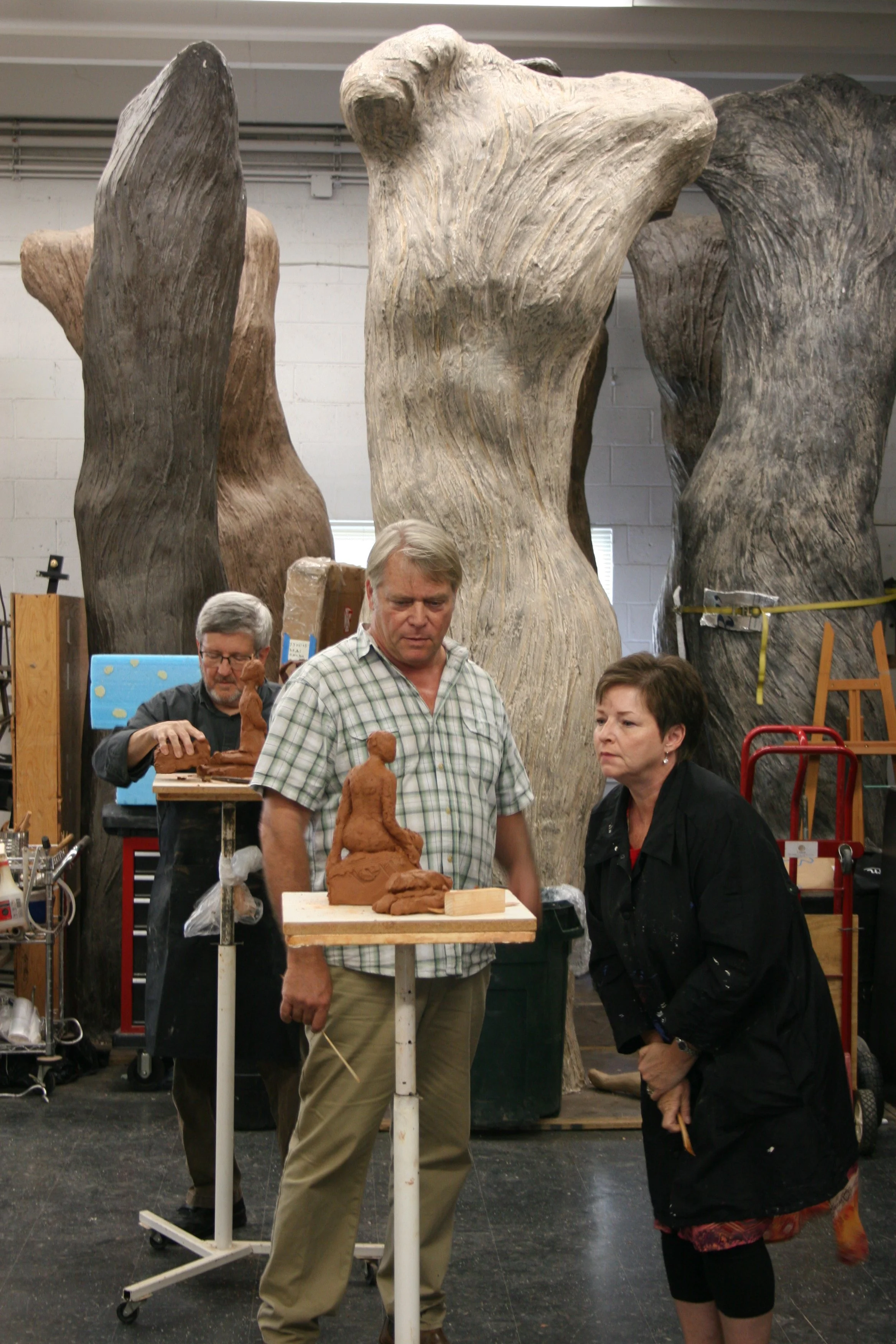 A group of people observing clay sculptures in an indoor art studio. Behind them are large wooden sculptures.