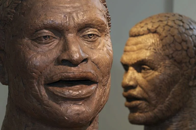 Two clay sculptures of male faces, one in the foreground with detailed features and open mouth, and the other in the background with textured skin.  Cultural Heroes