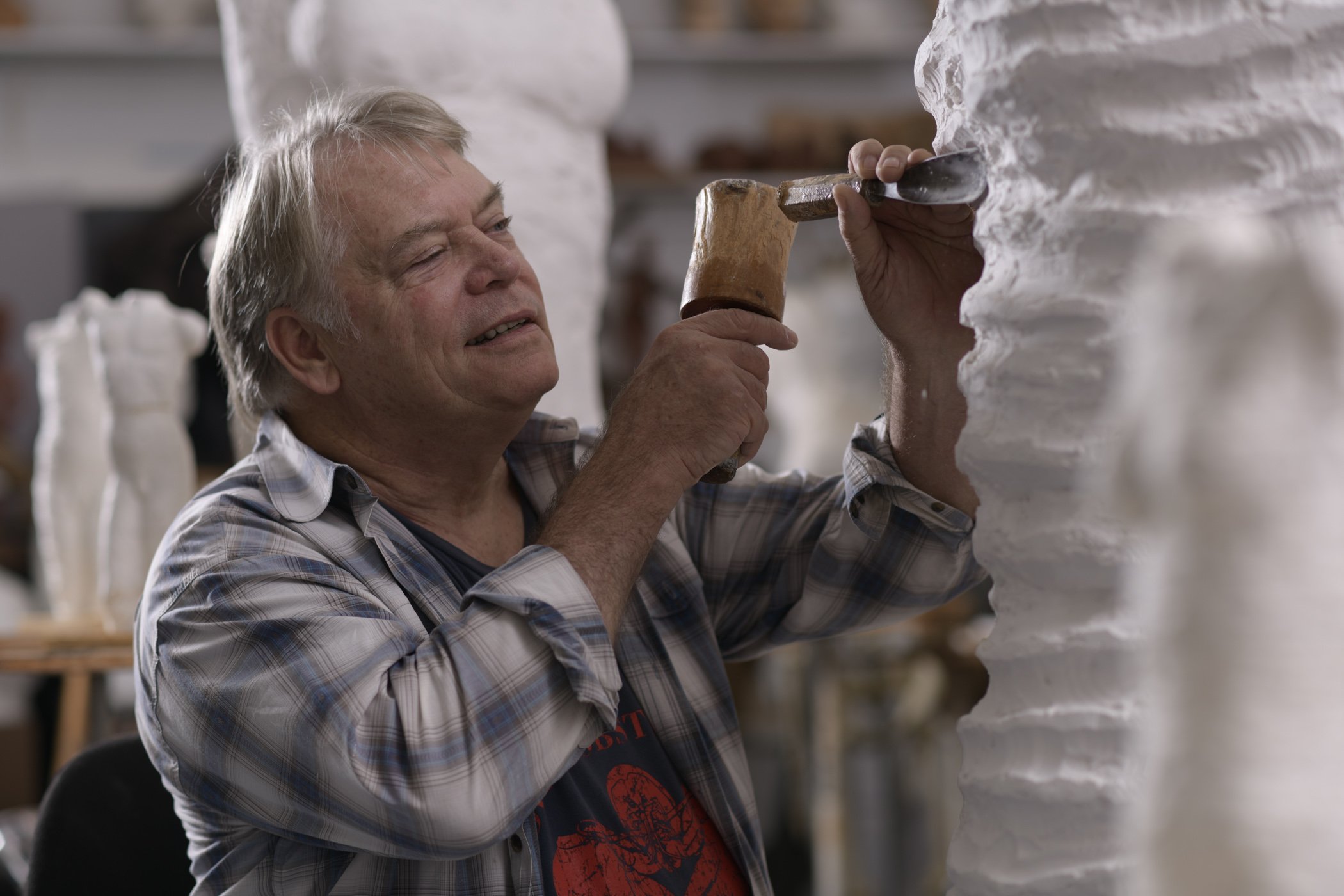 An older man with gray hair wearing a plaid shirt is holding a chisel and a hammer, carving or shaping a large white sculpture in a workshop.